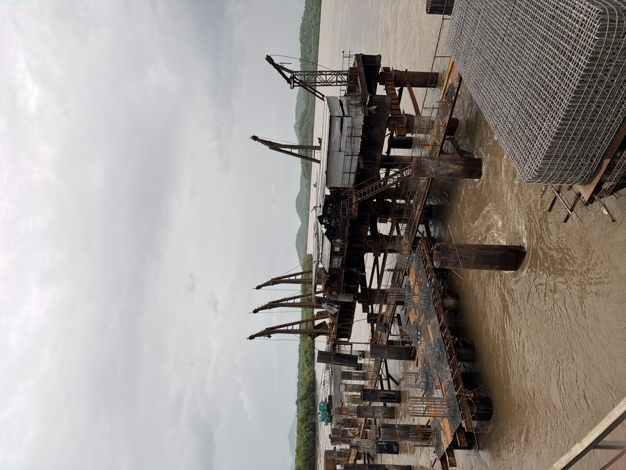 The image depicts a construction site in Vasai-Virar, India, likely on the Vasai Creek, a tidal estuary. A large, partially constructed bridge or pier structure dominates the scene, extending into the murky, brown water. The structure is supported by numerous large, rusty, cylindrical pillars. On top of the central platform of the structure, there are several industrial cranes, some appearing old and weathered, with visible cables and mechanical parts. A corrugated metal shed or building is also present on the platform.
In the foreground, to the right, is a large, rectangular cage-like structure filled with metal rebar, suggesting ongoing construction. To the left, more support pillars and scaffolding are visible, with metal rods protruding from concrete bases. In the middle ground, on the water, a turquoise construction vehicle with an extended boom, possibly a crane or excavator, is positioned on a barge or pontoon, actively engaged in work.
The background shows a lush, green, tree-covered coastline with hills in the distance under a heavily overcast, grey sky. The water is choppy, indicating a tidal current or wind. No people are clearly visible in the frame, but the presence of construction equipment and materials strongly implies human activity related to the project. The overall impression is one of an active, large-scale infrastructure project in progress, surrounded by a natural, watery environment.