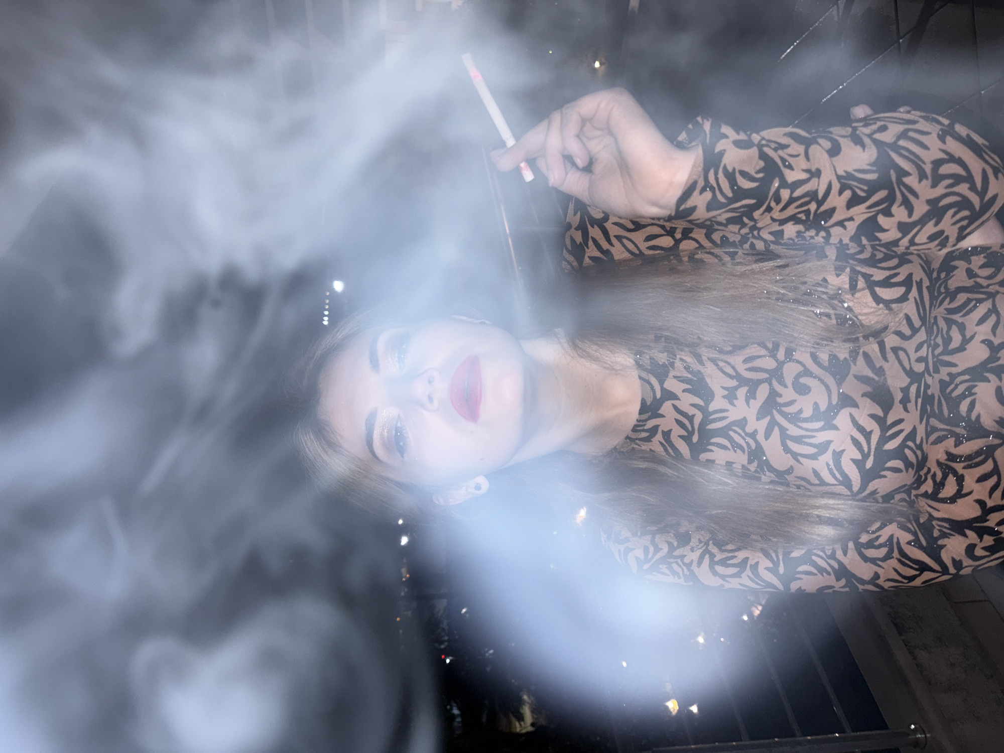An individual, identified as female, is depicted in a chest-up, portrait-style photograph taken at night in Budva, Montenegro. She possesses long, light brown hair, wears red lipstick, and has visible eye makeup. Her attire consists of a long-sleeved garment featuring a light brown or beige base with a dark, intricate, leaf-like pattern and subtle shimmering elements. The subject holds a lit cigarette, identifiable by a pink band near the filter, between her right index and middle fingers. She looks upwards towards the camera. Dense white smoke, likely originating from the cigarette, envelops her head and upper torso, illuminated by a frontal light source that also produces a noticeable lens flare. The background is dark, revealing numerous out-of-focus city lights and blurry landscape elements, viewed from an elevated position. A metallic railing is partially visible on both sides of the subject.