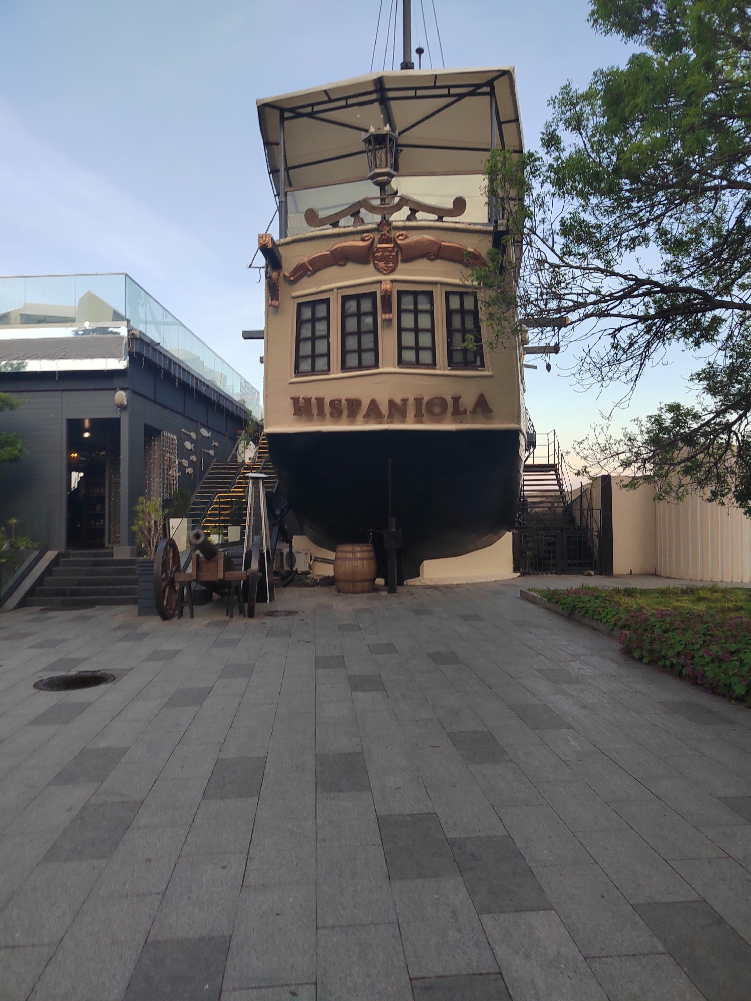 Here is a detailed description of the image:
The image captures a peculiar architectural structure in an urban setting. At the center stands a building designed to resemble the stern of a ship, complete with windows, and the name "HISPANIOLA" emblazoned across it. This ship-like structure is positioned between two buildings, giving the impression that it has been beached or deliberately placed in this location.
To the left of the ship, there is a modern building with a dark facade, featuring a restaurant or shop, indicated by the open entrance. A glass-enclosed area tops the building. Adjacent to the ship on the right is a low white wall, set in a garden area filled with greenery and flowers.
The foreground presents a wide, paved pedestrian area, which occupies a substantial portion of the frame. The paving stones are arranged in a rectangular pattern, directing the viewer's gaze toward the ship. A manhole cover is visible on the pavement.
Several objects are placed around the ship. These include a cannon, which adds to the nautical theme, a barrel, and a stepladder that leans against the side of the ship. Staircases are positioned on either side of the ship-shaped building, providing access to the upper levels.
To the right of the ship-like structure is a mature tree.
The image is shot during daylight, and the lighting suggests late afternoon or early evening. The location of this scene is not discernible from the image but might be a tourist attraction or an area with a unique architectural theme.
