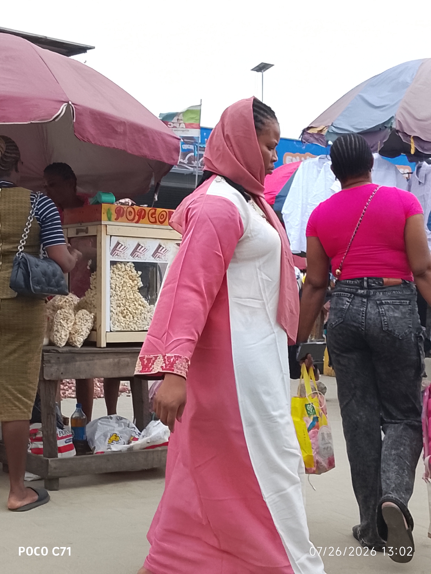 The image depicts an outdoor market scene in Lekki, Nigeria. In the foreground, a woman wearing a pink and white abaya walks past a popcorn stand. The popcorn stand is illuminated and filled with popcorn. To the left, another woman wearing a green dress and a blue and white striped shirt is partially visible, holding a dark handbag. In the background, various stalls are set up, displaying clothing and other goods, with awnings providing shade. The ground is paved, and there are people milling about in the distance. The overall atmosphere suggests a bustling marketplace.
