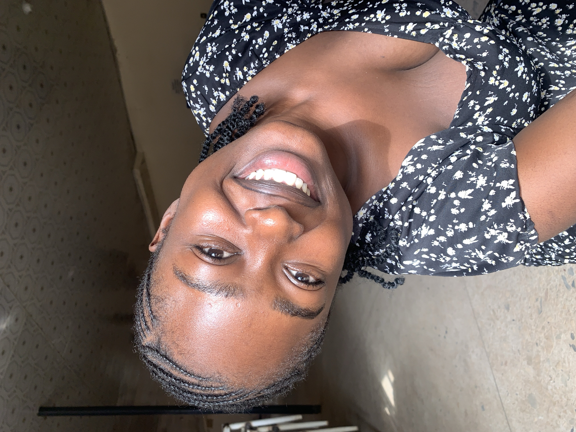 This media file features a close-up portrait of a young person with dark skin, smiling widely directly at the camera. The individual's head is tilted back, and they appear joyful and content. They have dark eyes, neatly groomed eyebrows, and intricate cornrows or braids styled along their hairline and sides. Short facial hair, resembling stubble or a closely trimmed beard, is visible around the chin and sideburns. Their bright, broad smile reveals healthy white teeth and slightly glossy lips.
The person is wearing a black top adorned with a pattern of small white and subtle yellow flowers. The fabric appears light.
The background suggests an indoor or semi-covered outdoor location, consistent with "New Stadium, Nigeria." On the upper left side of the frame (when viewed upright), there is a wall with a subtle, dark geometric pattern. The lower right side reveals a light-colored, speckled floor, possibly terrazzo or concrete, showing reflections from strong natural light. A dark, thin horizontal bar-like object is present near the bottom of the image. The bright illumination indicates it is daytime. No visible text is present.