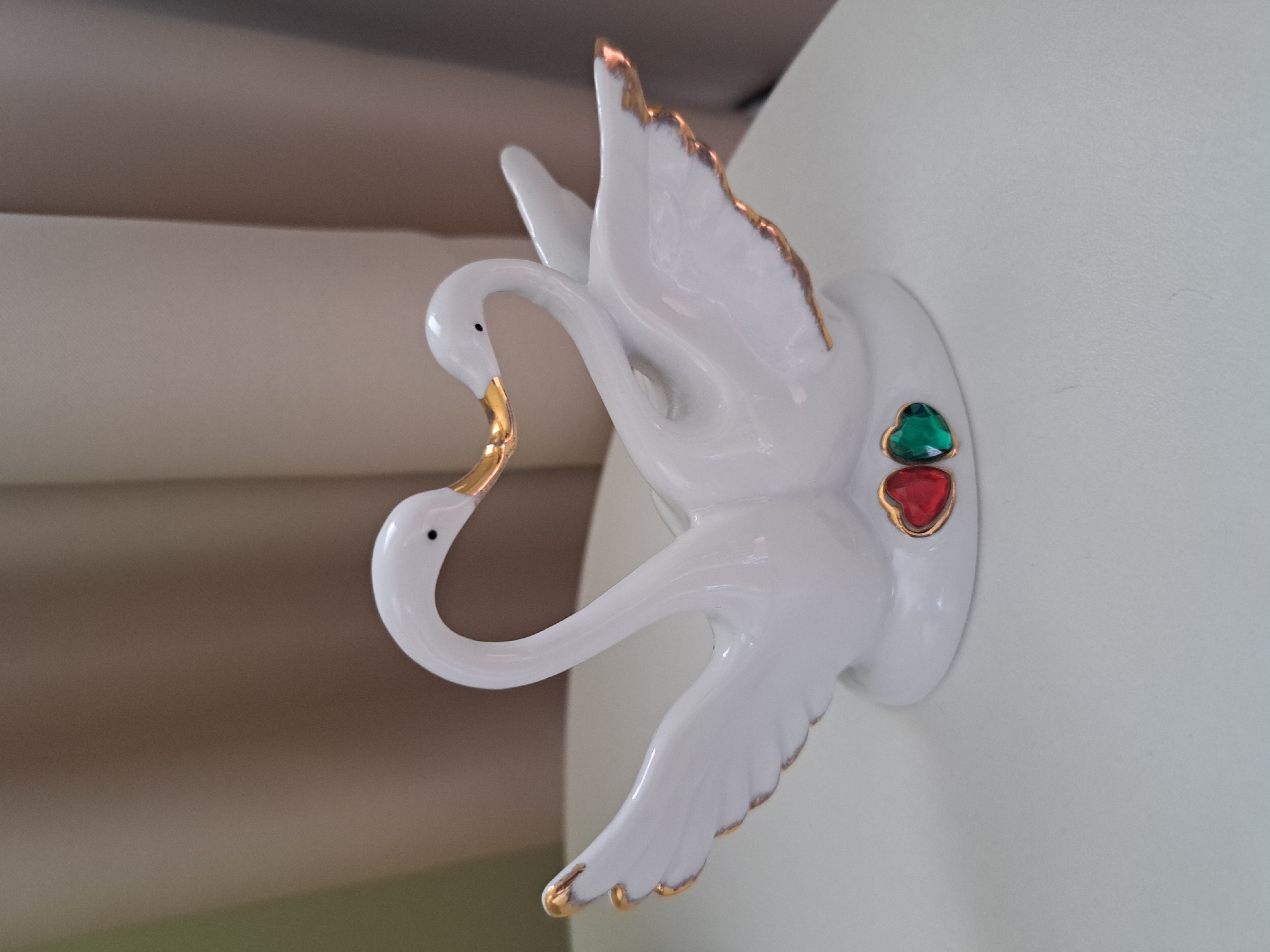 A white ceramic figurine of two swans facing each other with their necks forming a heart shape is centered on a white surface. The swans have gold accents on their beaks and wingtips. Two heart-shaped gemstones, one red and one green, are affixed to the base of the figurine. The background consists of soft, beige curtains. The scene is indoors, and no specific location within Irkutsk, Russia, can be determined from the image.