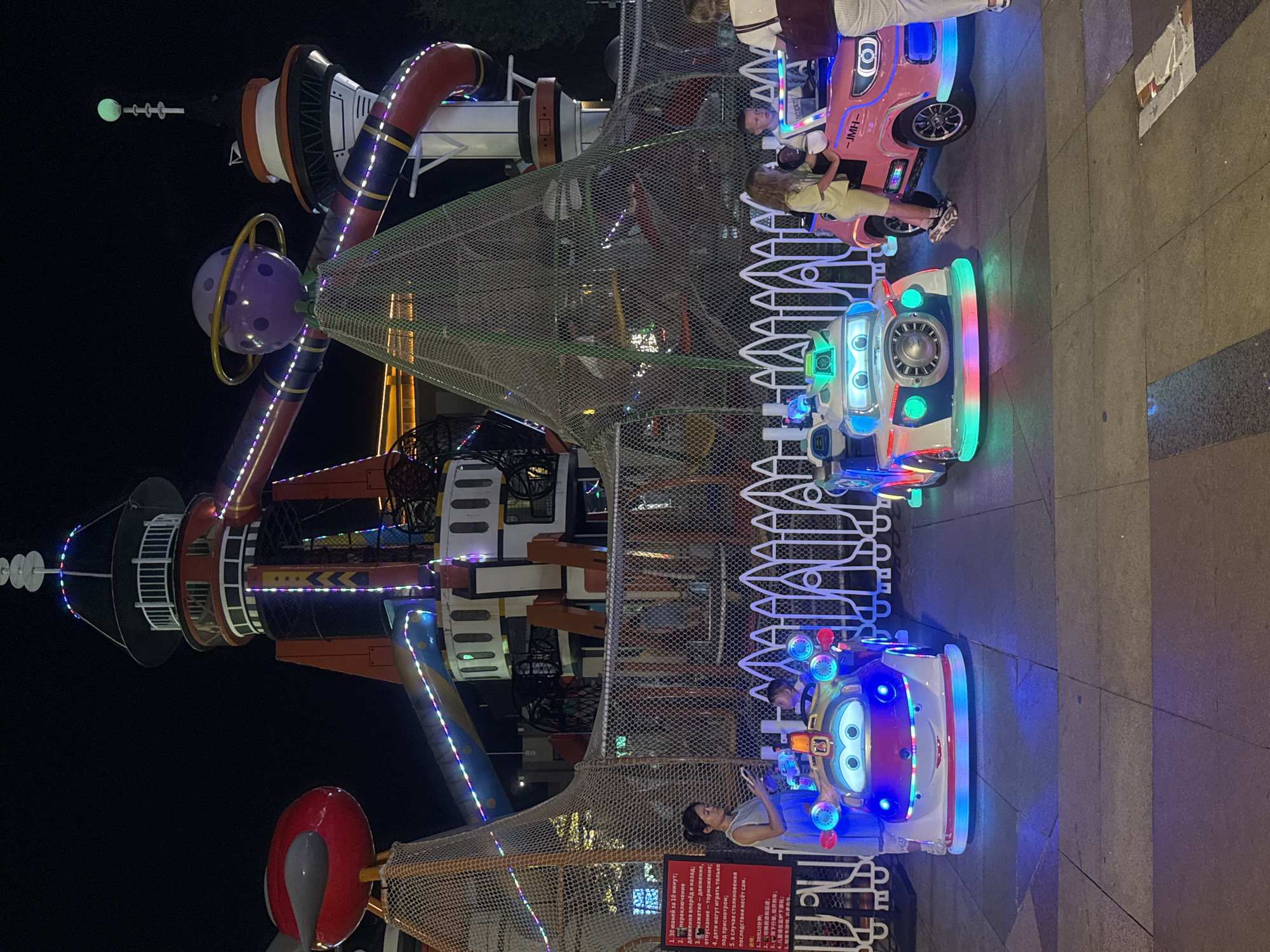 The image captures a nighttime scene at an amusement park, specifically in the city of Qinhuangdao, China. In the foreground, three brightly lit, cartoon-themed electric cars are parked on a paved surface. The car on the left is white and blue with large, expressive eyes and colorful flashing lights. To its right is a pink car with similar cartoon features, and it has "JMH" written on its side. A young girl is standing next to the pink car, and a boy is seated inside it. To the far right, a woman in white pants and a dark bag is partially visible. A woman in a light blue dress is standing near the white and blue car, holding a phone. In the background, a large, futuristic-looking amusement ride structure dominates the scene, featuring colorful lights, slides, and what appears to be a rocket-like tower. A red, egg-shaped object is suspended on the left side of the ride. A net covers a portion of the structure in the middle. A red sign with Chinese text is posted to the left of the electric cars, detailing ride rules or information. The overall atmosphere is festive and playful, illuminated by the colorful lights of the rides and the electric cars.