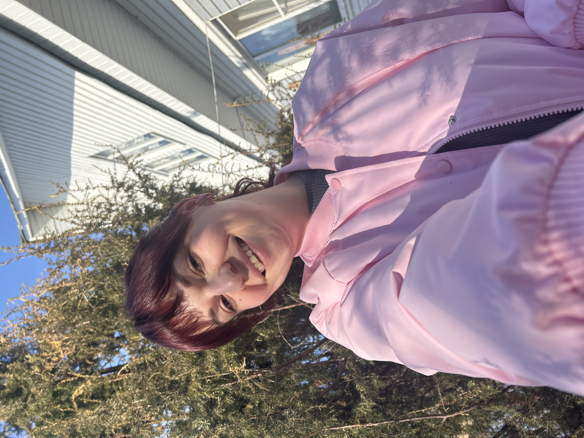 A medium shot depicts a woman wearing a pink jacket and smiling at the camera. Her auburn hair with bangs is visible. The background includes a white, modern building with horizontally-oriented siding and a large window. Behind the woman, a coniferous tree with bare branches fills a portion of the frame. The sky is visible in the upper left corner. The photograph appears to be taken from a low angle. There is no information to determine the precise location within the city of Фоки, Russia.