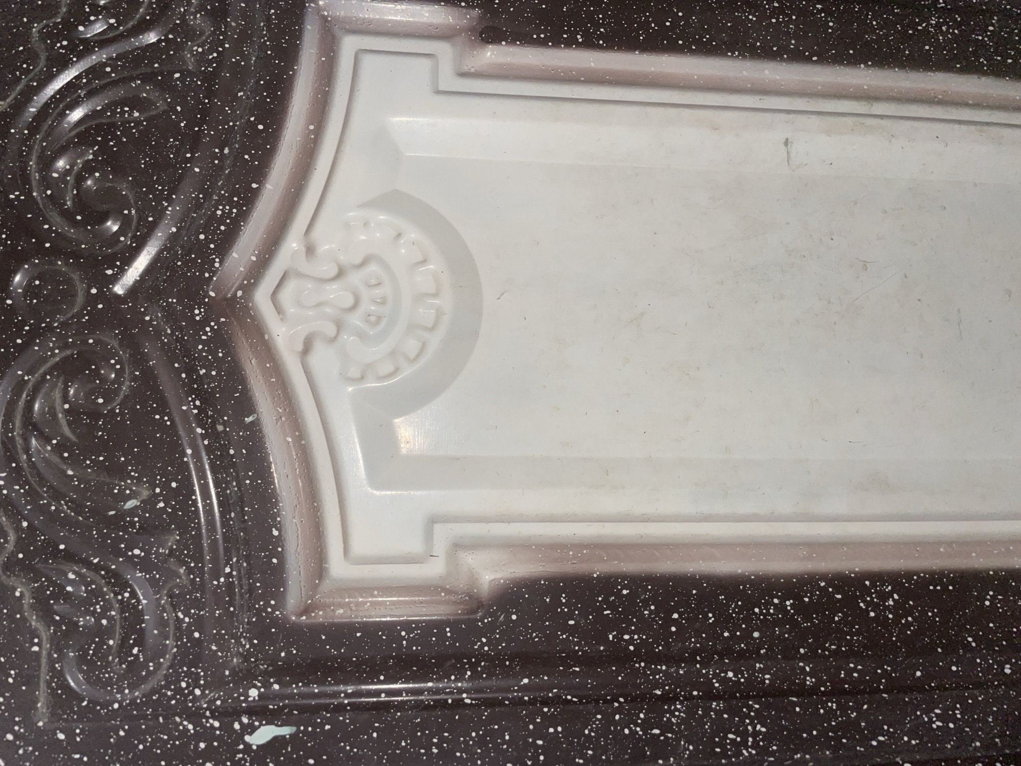 The image displays a close-up view of a dark brown, speckled surface with a white, recessed panel in the center. The dark brown area has a textured, possibly painted, finish with small white speckles scattered across it. Elaborate, raised decorative swirls are visible in the upper part of the dark brown section. The white panel is bordered by a slightly raised, light brown edge. The white panel itself appears to be made of a smooth, matte material and features a decorative embossed element at the top. The scene is indoors, with no visible environmental context like a city, or specific location in Jalingo, Nigeria. There are no people, actions, or interactions depicted.