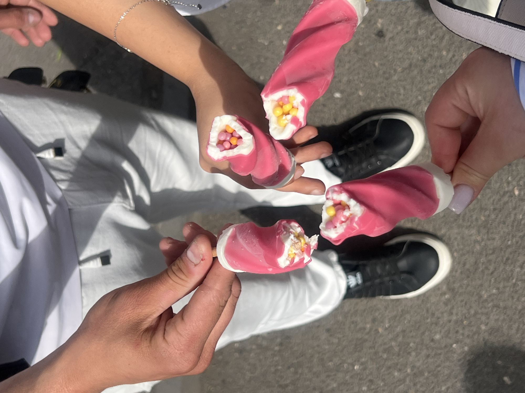 The image shows a group of people holding pink and white twisted ice cream pops with colorful candy centers. The people are dressed in light-colored clothing. The ground appears to be paved. Parts of black and white sneakers are visible. Yerevan, Armenia is not discernable from the image.