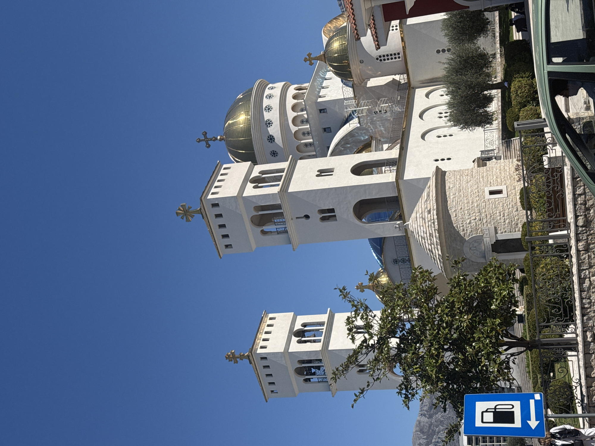 The image shows the Serbian Orthodox Church of St. John Vladimir in Bar, Montenegro. The building features white walls, multiple golden domes, and two prominent bell towers topped with crosses. A small tree is located in front of one of the towers, partially obscuring it. There is a blue road sign with a luggage icon and a downward arrow in the bottom right corner. The background is a clear blue sky. Green trees and parts of a fence are visible to the side of the church. The image is oriented vertically.