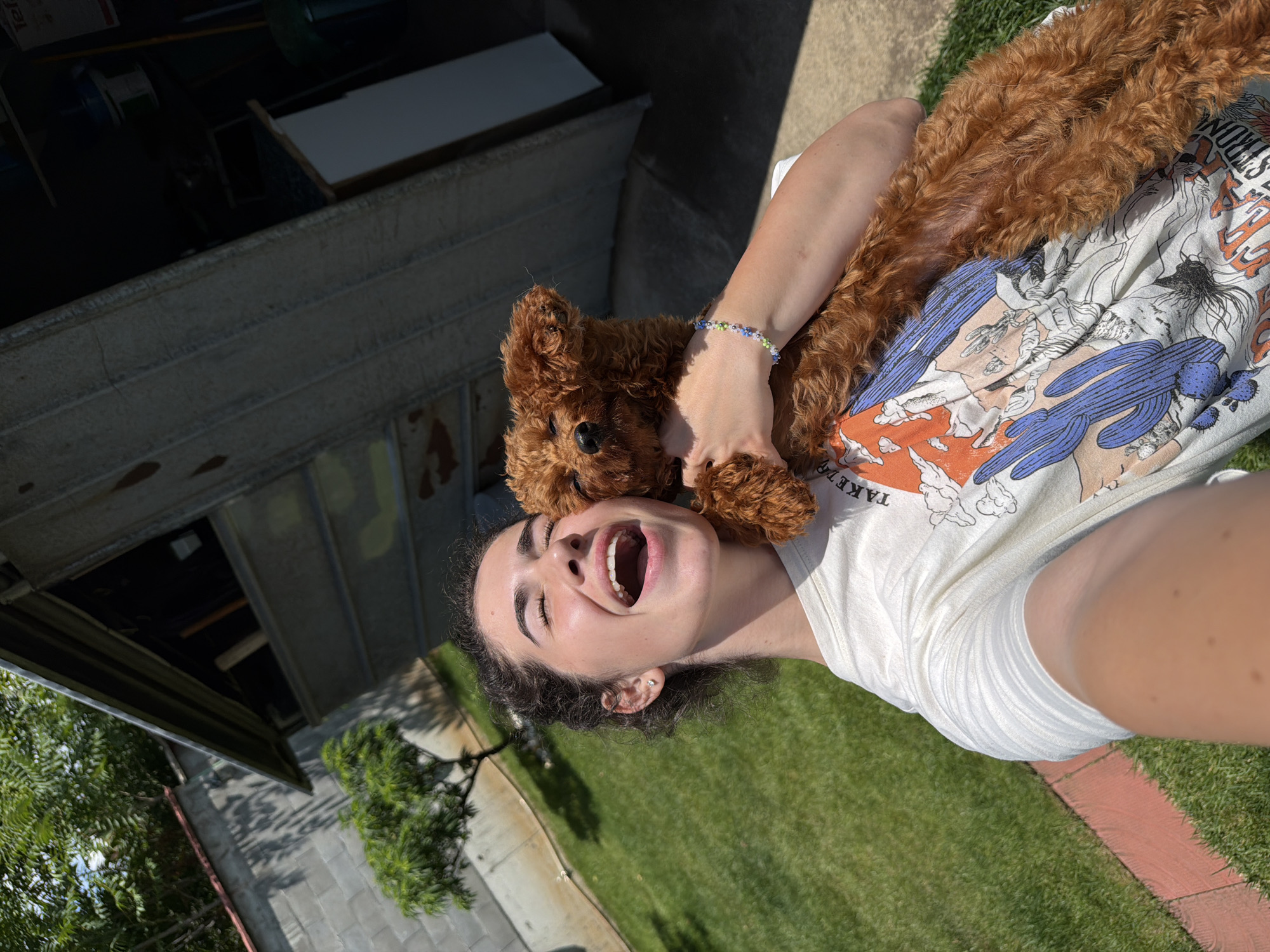 A young person with dark curly hair is lying on a lawn, holding a small, fluffy, reddish-brown dog. The person is wearing a white t-shirt with a graphic print featuring cacti and desert animals. Their mouth is wide open in a laugh, eyes closed, and they have on an earring. The dog is resting on the person's chest and neck, with its head near the person's face. A beaded bracelet with green, blue, and white beads is visible on the person's wrist. The scene is outdoors, with a grassy lawn in the foreground and a concrete structure with an open compartment in the background. Sunlight suggests it is daytime. The overall impression is one of joy and affection between the person and the dog. The setting appears to be a backyard in Sofia, Bulgaria.