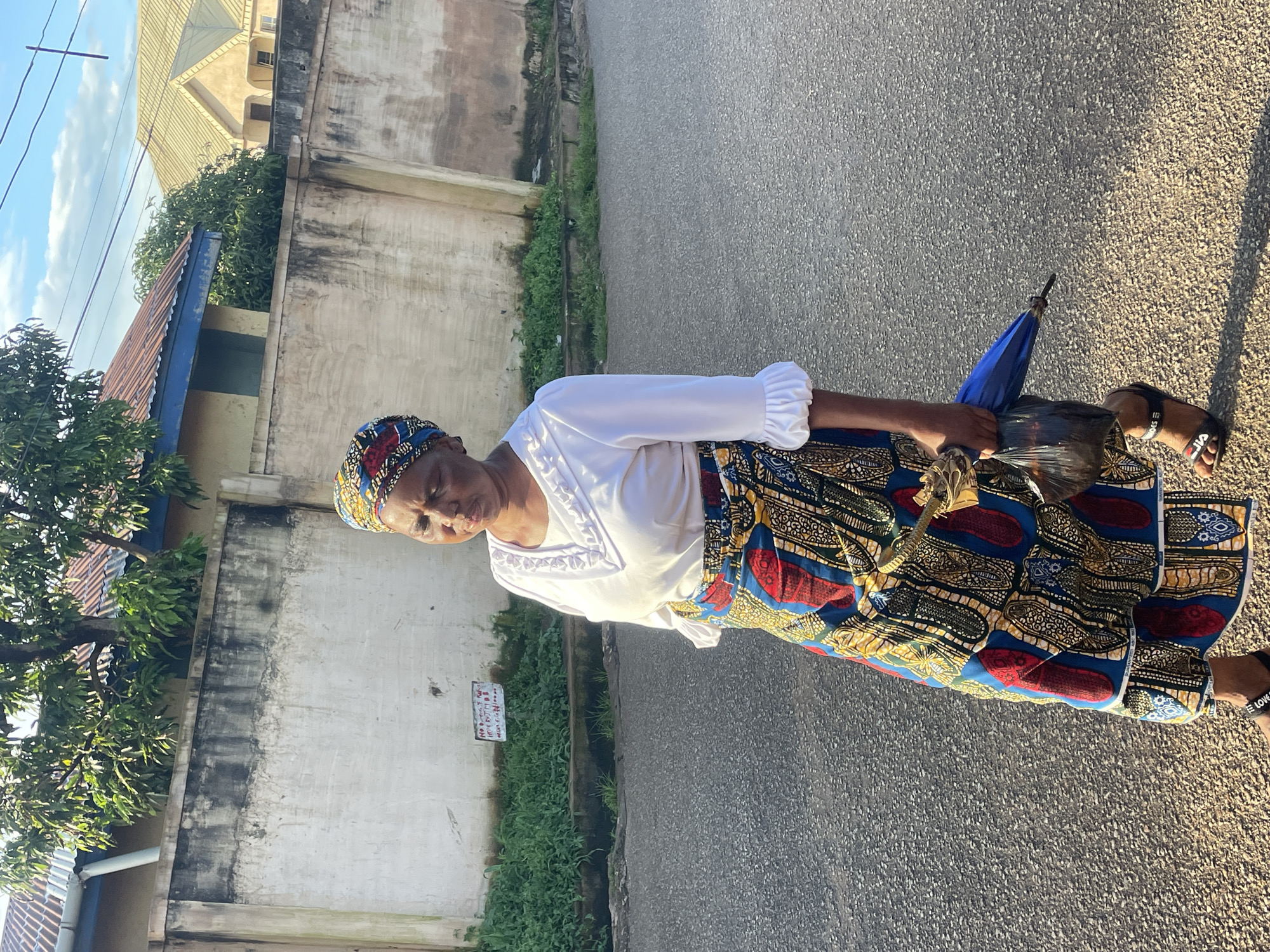 A middle-aged woman, dressed in a vibrantly patterned Ankara skirt and a white blouse with ruffled sleeves, walks on a paved road in Jos, Nigeria. She is holding a dark plastic bag containing a blue umbrella in her right hand. The woman wears black sandals with white lettering. Behind her, a white concrete wall runs along the side of the road, with a strip of overgrown green vegetation bordering it. Beyond the wall, glimpses of buildings with tiled roofs are visible. The sky above is a clear blue with scattered clouds. The scene is captured in natural daylight, with shadows indicating the direction of the sun.