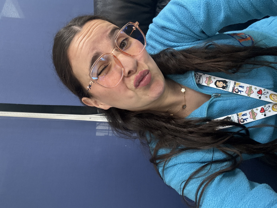 A female subject with dark brown hair, wearing large light-colored eyeglasses with prominent frames, is depicted in a close-up, chest-up shot. She is winking with her right eye and has her lips puckered. She wears a light blue fleece zip-up jacket and a gold-colored chain necklace featuring three clover-shaped charms, two dark and one light. A white lanyard with various colorful cartoon illustrations, including faces, hearts, stethoscopes, and text such as "I'm a Nurse," "Callie," "Dance It Out," and "MCSTEPMOM," hangs around her neck. The background consists of a solid dark blue surface, likely a wall or partition, with a vertical white beaded blind pull chain visible on the right side of the frame. A portion of a black object, possibly a chair back, is visible on the far right. The image contains no visual information to determine its specific location within the city of Meknes, Morocco, or any other geographical location.