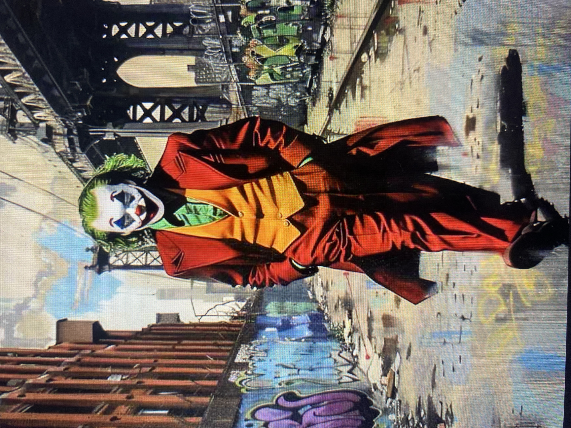 A digital illustration depicts the Joker character from the movie "Joker" walking down a wet, graffiti-covered alleyway in an urban setting. The Joker is wearing his signature red suit, orange vest, and green shirt, with his face painted in his iconic clown makeup. In the background, a bridge with intricate metalwork spans across the cityscape. The alley is lined with brick buildings on the left and a chain-link fence with more graffiti on the right. Puddles of water reflect the surrounding environment, and debris is scattered on the ground, contributing to a gritty and desolate atmosphere. The sky above is a mix of blue and white clouds.