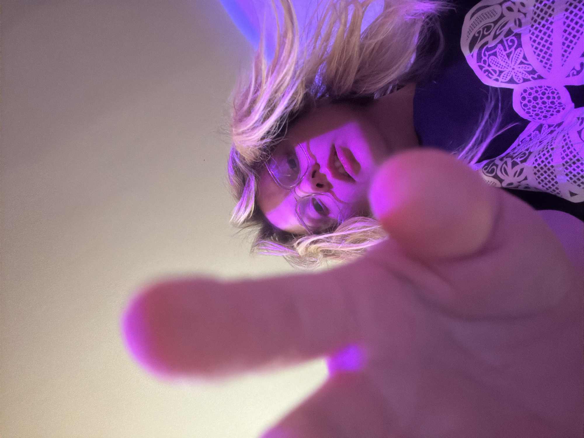 The image is a low-angle shot of a person with blonde hair, wearing glasses, and reaching towards the camera with their hand. The person is wearing a dark-colored garment with a white floral pattern near the top right corner of the frame. The image is lit with a strong purple or blue light. The background is a light grey color, but no specific location is discernible. The image does not provide any information to indicate the location is Tel-Aviv, Israel.