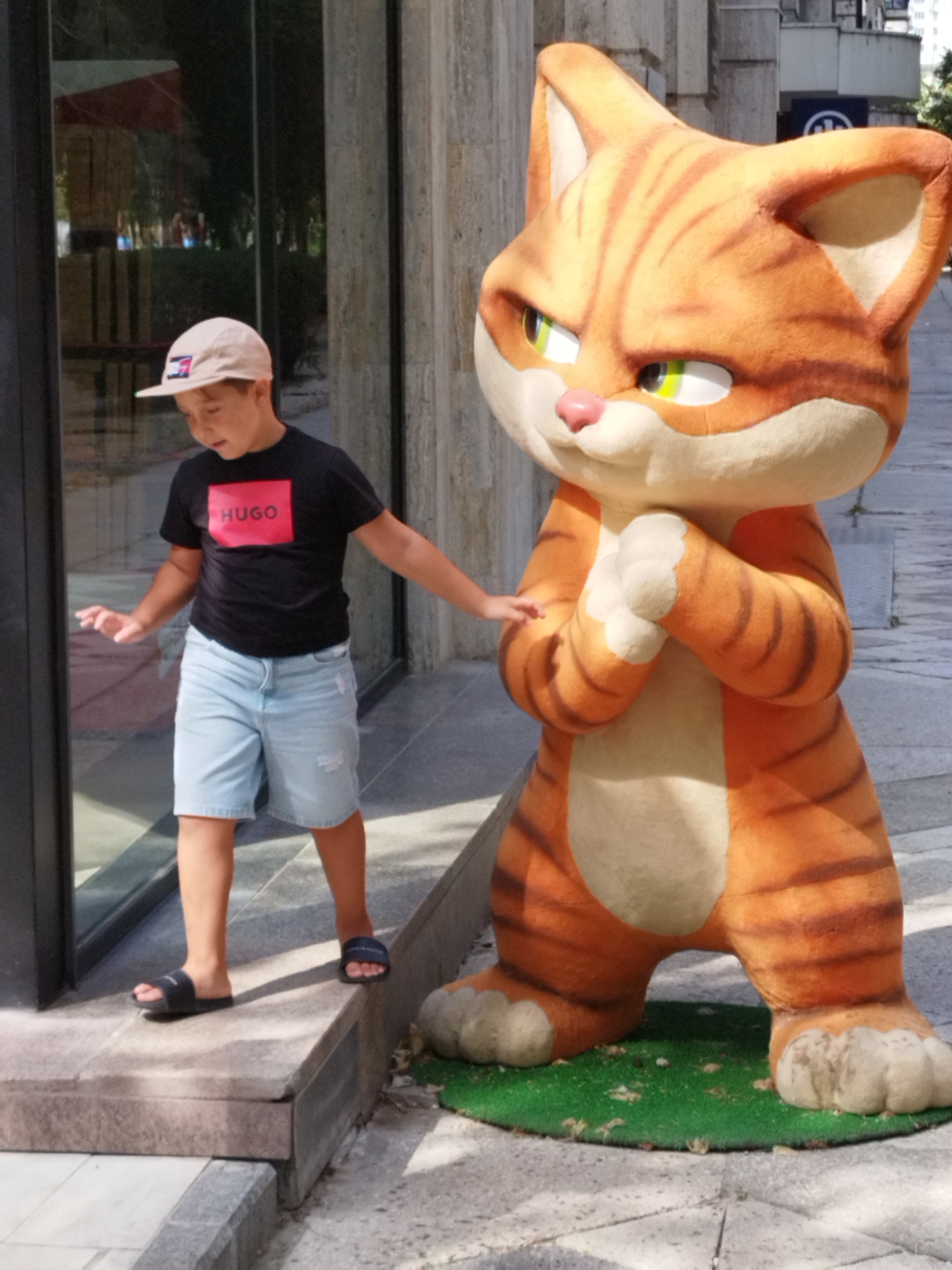 A young boy wearing a black t-shirt with a red "HUGO" logo, light blue denim shorts, a beige baseball cap, and dark blue slide sandals stands on a raised concrete platform. He is reaching towards a large, orange, cartoonish cat statue with green eyes and striped fur. The statue is positioned on a patch of green artificial turf next to the platform. The boy is positioned in front of a building with large glass windows and a stone facade. The scene appears to be outdoors in an urban environment, possibly a city street or pedestrian area, with paving stones visible in the background. The setting is consistent with locations in Bucharest, Romania.