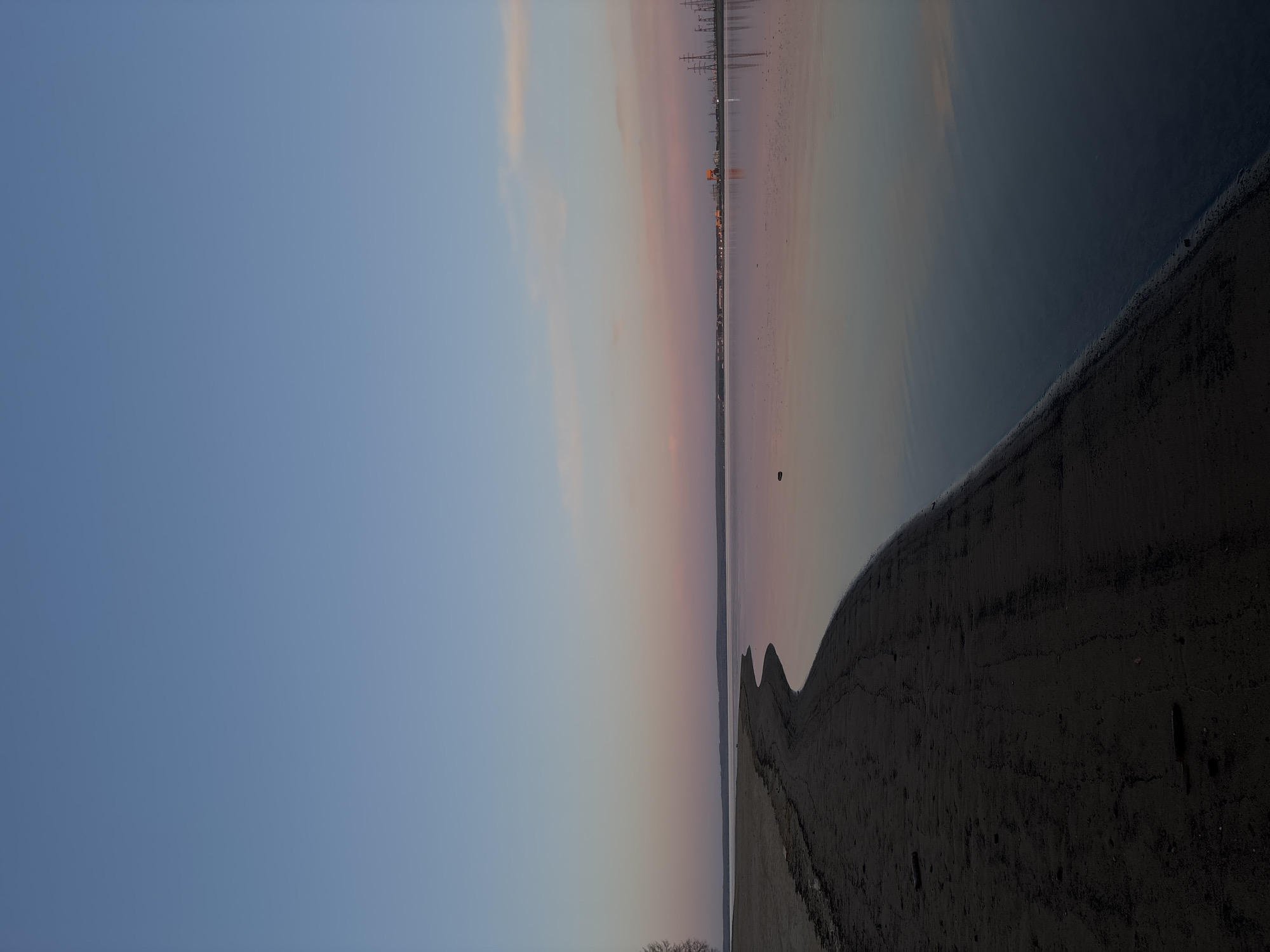 The image depicts a coastal landscape in Noviy, Russia at dusk or dawn. The sky transitions from a pale blue on the left to a gradient of pinks and oranges towards the horizon. Reflections of industrial structures, including power lines and buildings, are visible in the water. A dark, sandy shoreline curves along the right side of the frame. The horizon is straight, and the perspective is at eye level.