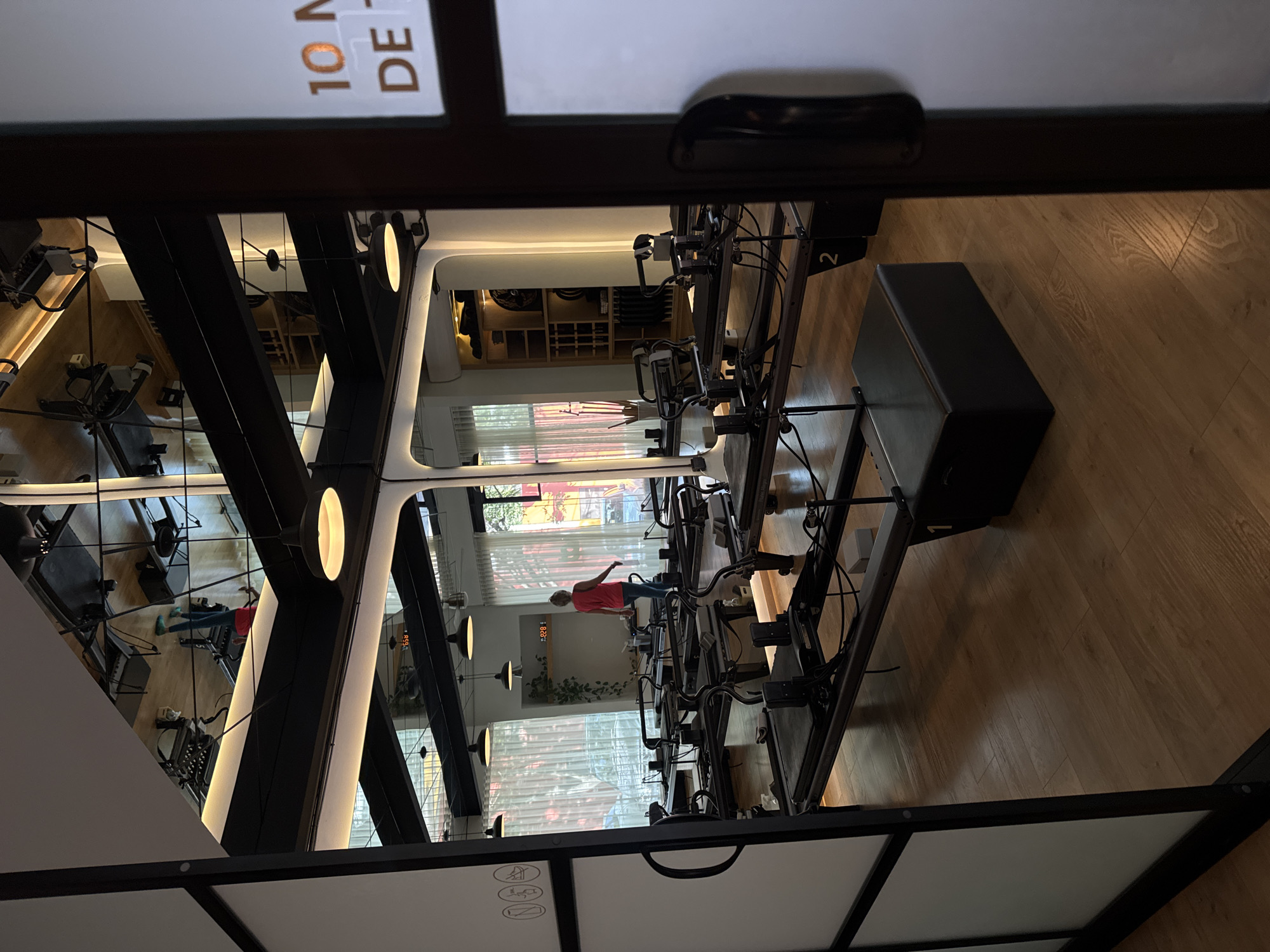 The image depicts a modern and well-lit Pilates studio, viewed from a slightly elevated perspective. A row of multiple black Pilates reformer machines extends from the foreground towards the back of the room. The studio features light-colored hardwood flooring and large windows with sheer white curtains lining one side, allowing natural light to illuminate the space.
In the mid-ground, a person wearing a red top and dark leggings is visible standing on one of the Pilates reformers, appearing to be engaged in an exercise or demonstration. The studio's aesthetic is contemporary, characterized by minimalist black metal beams, white walls, and various modern lighting fixtures, including pendant lights and track lighting. Mirrors on the opposite wall reflect the room, creating an illusion of expanded space and offering additional views of the equipment and the environment.
Notable details include a dark rectangular box or bench on the floor to the right, and shelving units with various items visible in the background. On a white surface in the foreground, partial text "10 M" and "DE" is visible. Additionally, a number "2" is seen on one of the machines, and reflections in the mirrors show numerical displays resembling clocks or timers, reading "12:21" and "9:59". The scene conveys a calm and organized fitness environment, likely situated in Puebla, Mexico.