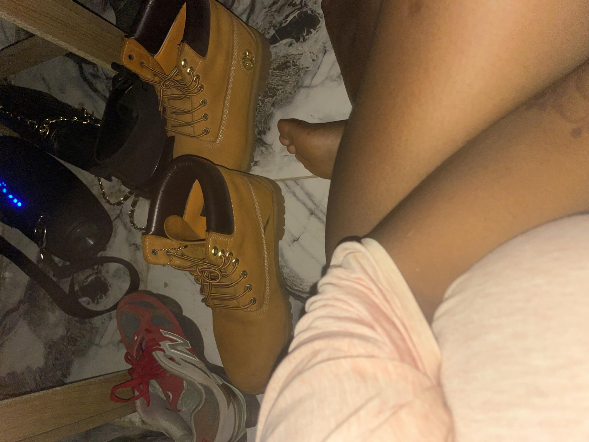 The image shows a pair of tan Timberland boots and a red and gray New Balance sneaker on a marble-patterned floor. A person's legs, clad in a pink garment, are visible in the foreground. In the background, a black bag with a gold chain and a black speaker with blue lights are present. The scene appears to be indoors, possibly in a residential setting, and is not clearly identifiable as being in Keffi, Nigeria, based solely on the visual information provided.