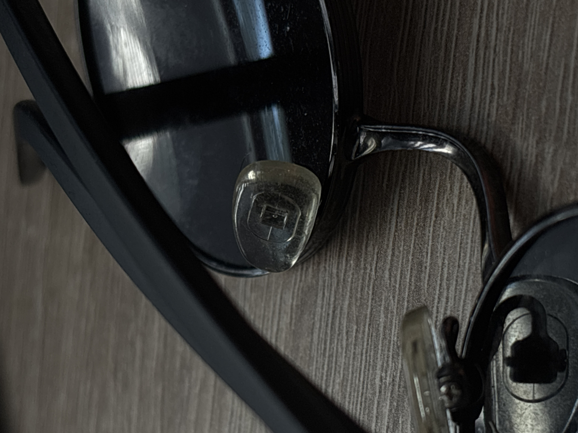 A close-up, eye-level shot shows a dark-rimmed pair of sunglasses with a clear nose pad, resting on a wooden surface. The sunglasses are positioned diagonally across the frame, with the left arm and part of the lens visible. The wooden surface has a visible grain pattern. No people are present in the image. The scene does not contain any specific details that would indicate a location within the city of Saint Petersburg, Russia.