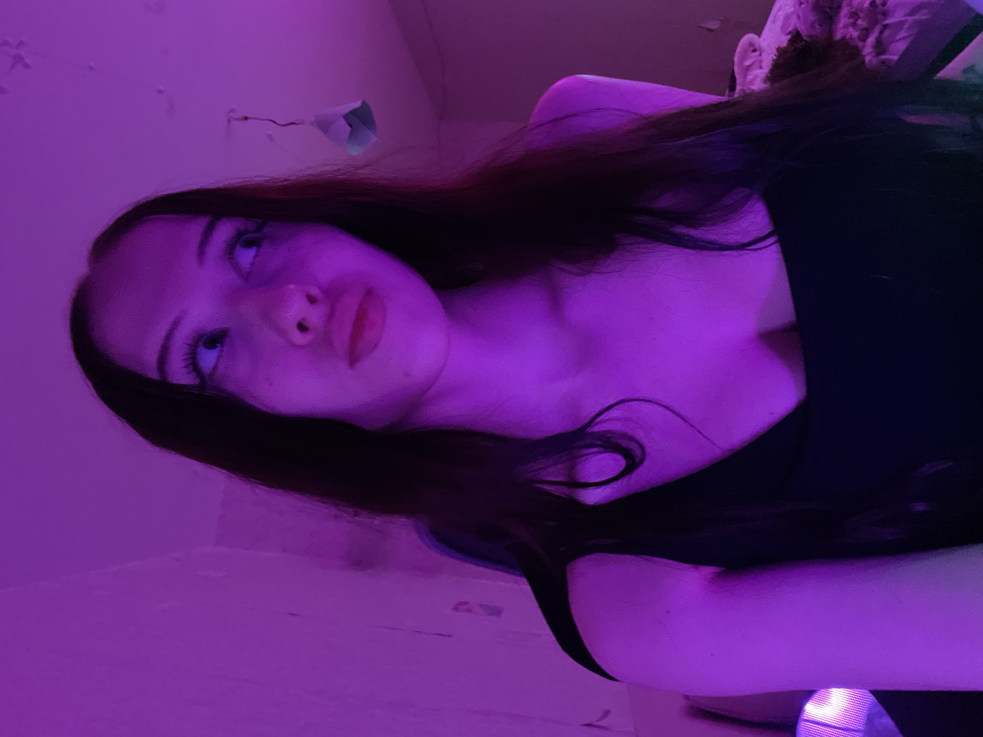 The image shows a young woman indoors, illuminated by purple lighting. She has long, dark hair and is wearing a black top. She is looking to her right, with her mouth slightly open. The background is a wall that appears to be painted a light color, with some texture visible. There are some objects hanging from the ceiling, including what looks like a small, folded piece of paper or fabric. To the left of the woman, a portion of a bed or couch is visible, with some bedding. The overall setting suggests a bedroom or living space. There is no indication of the specific location within Votkinsk, Russia.