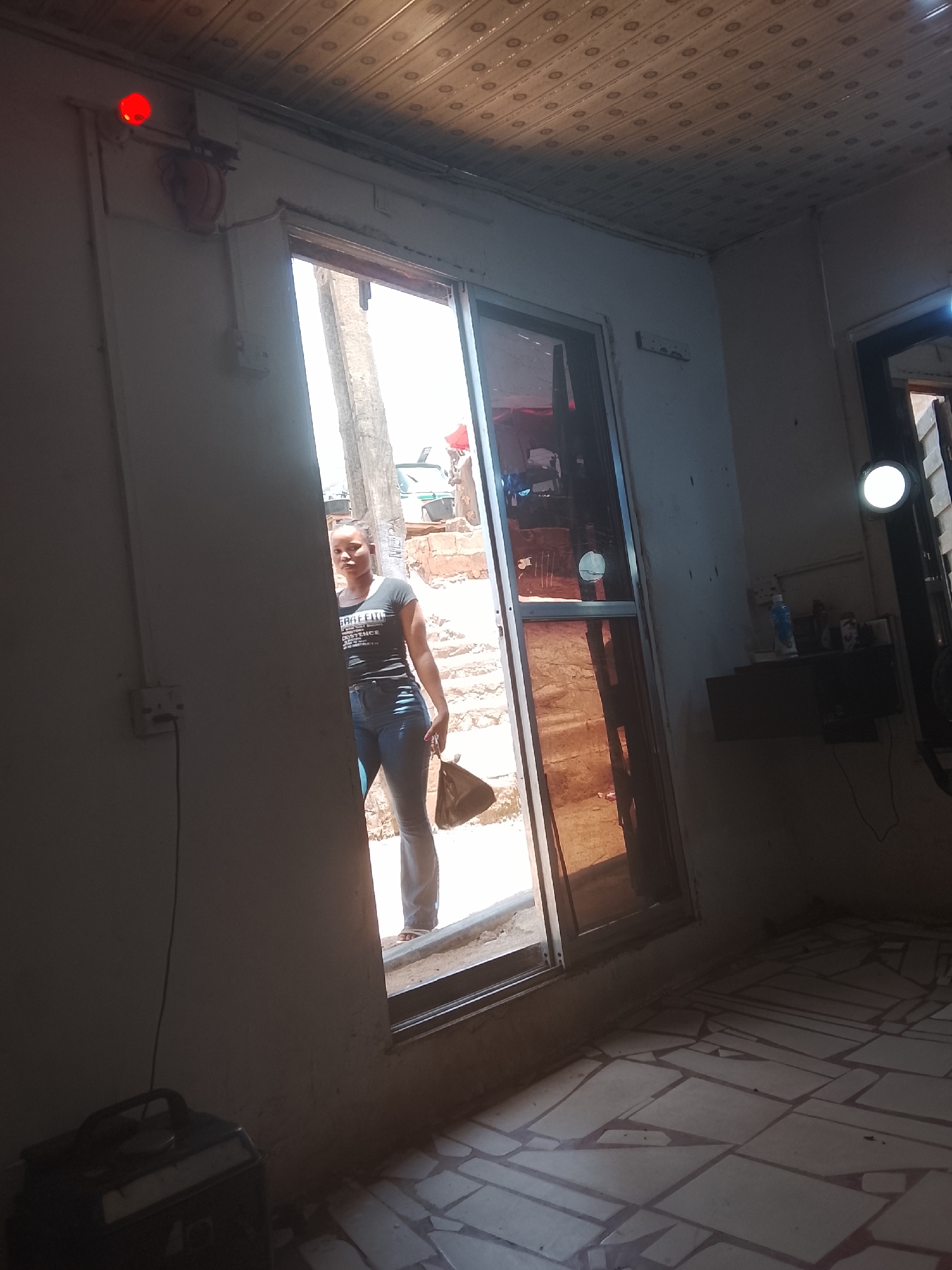 The image shows a woman standing in a doorway. She is wearing jeans and a t-shirt and is holding a purse. The doorway leads to an exterior location featuring concrete stairs. A red light is mounted on the wall above the door frame. The interior appears to be a room with a tiled floor and some household items on a shelf. The location is in the city of Unknown, Unknown.
