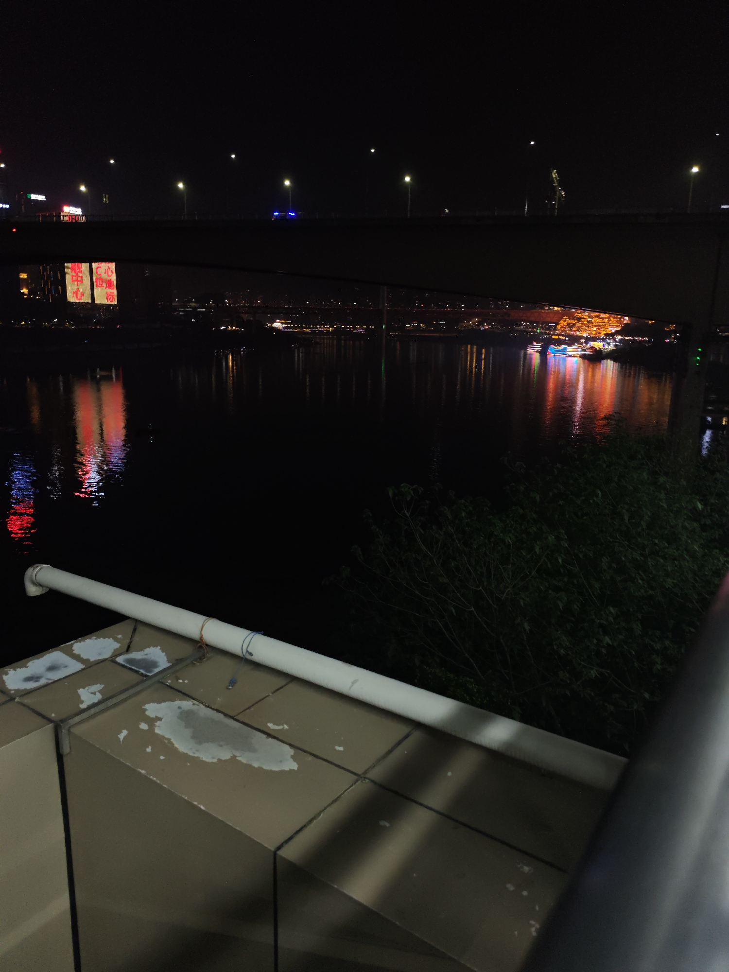 The image depicts a nighttime urban landscape, likely within the Yuzhong District of China, given the location tags. A body of water reflects city lights, with a bridge spanning the scene horizontally. The bridge features evenly spaced streetlights. Light reflects in the water. In the foreground are a wall with weathered tiles and a pipe. A bush is also visible.