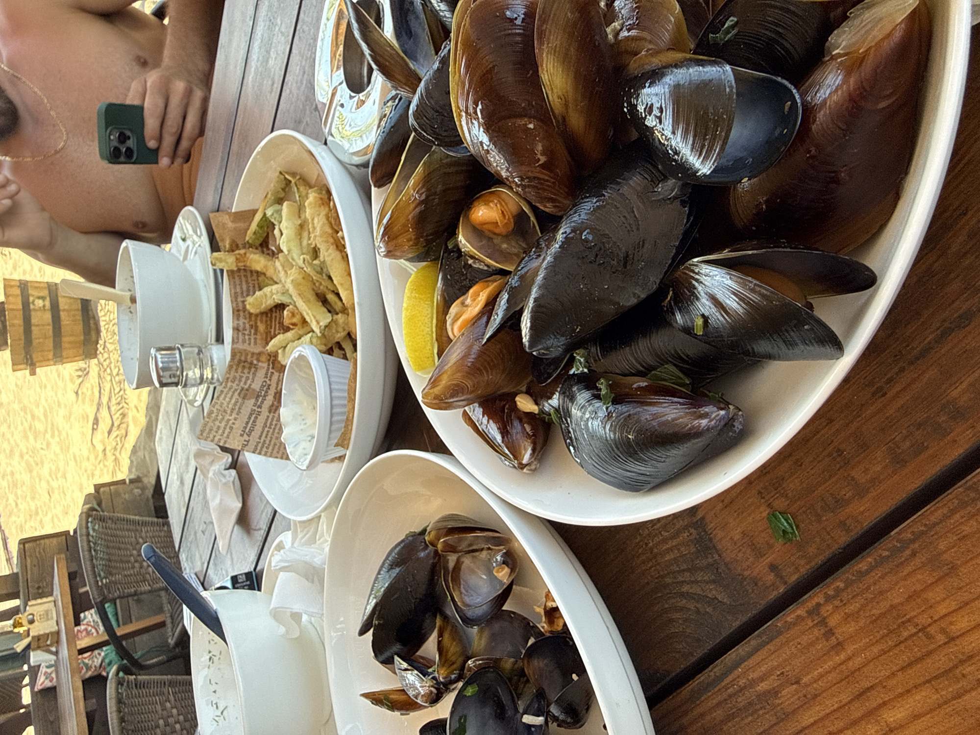 The image depicts a close-up of a wooden table at an outdoor restaurant, likely on the beach in Dyuni, Bulgaria, given the sandy background and beach umbrellas. Two white platters of mussels are the primary focus, with one platter in the foreground filled with dark, glossy mussels, some open revealing their orange flesh, and garnished with small green herbs. A lemon wedge is visible on this platter. To the left, a smaller white platter also holds mussels. Behind the mussels, a bowl of french fries is visible, along with a small dish of dip and some crumpled paper. In the background, a man is partially visible, shirtless, holding a smartphone. The overall atmosphere suggests a casual dining experience by the sea.