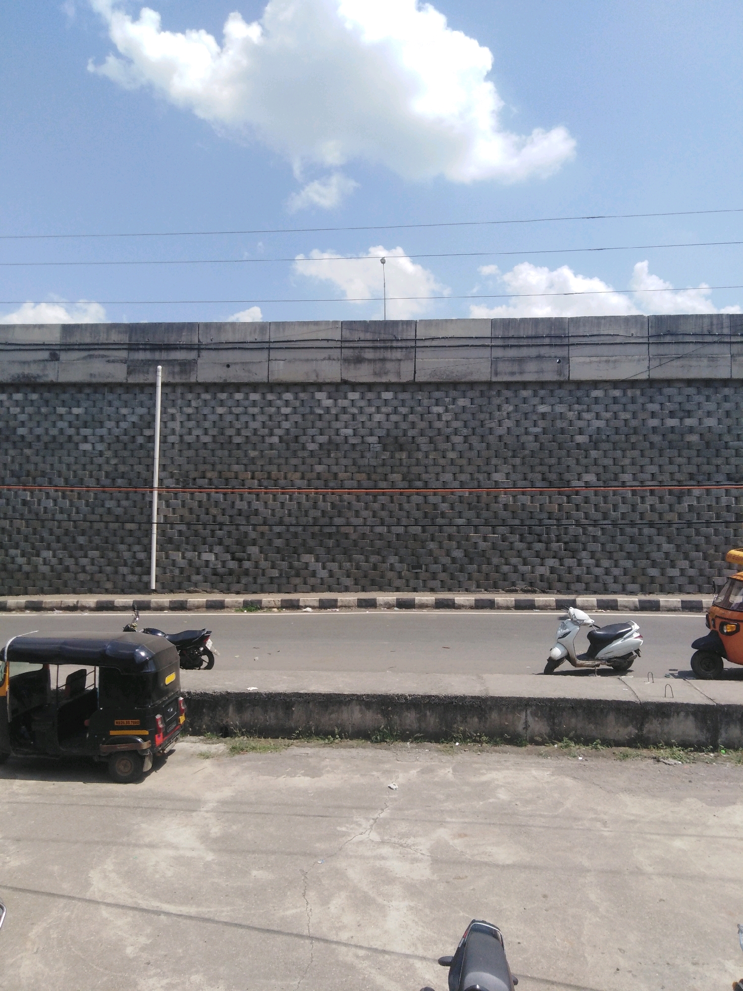 The image depicts a street scene in Nanded, India, under a clear blue sky with scattered white clouds. A large, textured concrete wall dominates the background, with electrical wires running along its top. Below the wall, a two-lane road is visible, separated by a concrete divider. Several vehicles are present: a black auto-rickshaw is parked on the left side of the road, and a white scooter and an orange auto-rickshaw are parked further down on the right. A dark motorcycle is also visible parked near the auto-rickshaw. The road is bordered by a concrete sidewalk and then a concrete ground surface in the foreground. The overall impression is an urban street with typical traffic elements.