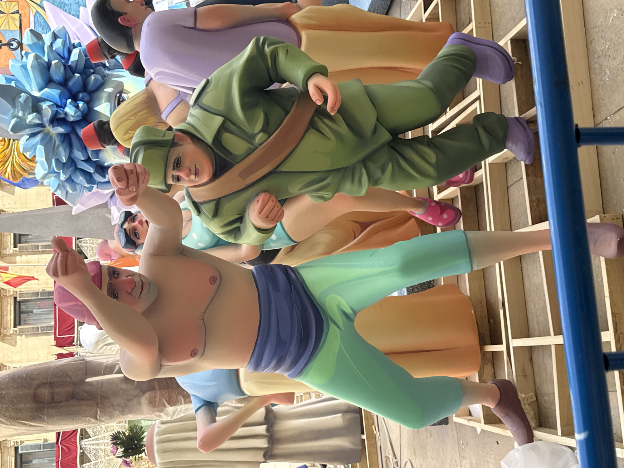 A scene depicts a group of brightly colored, large-scale figures, likely part of a festival or parade float, set up outdoors on a wooden platform. In the foreground, a figure with a pink hat and no shirt, wearing blue pants, is positioned on the left. To the right of this figure, a person in a green military-style uniform with a brown strap across their chest is running forward. In the background, other figures are visible, including one in a purple top and another with a blonde hairstyle. A large, abstract, blue, crystalline structure is prominent in the upper right portion of the image. The figures are rendered in a cartoonish style with bold colors and simplified features. The ground appears to be paved, and a blue railing runs across the bottom of the frame. In the background, a building with windows and a Spanish flag can be seen. The overall impression is one of a festive and artistic display.