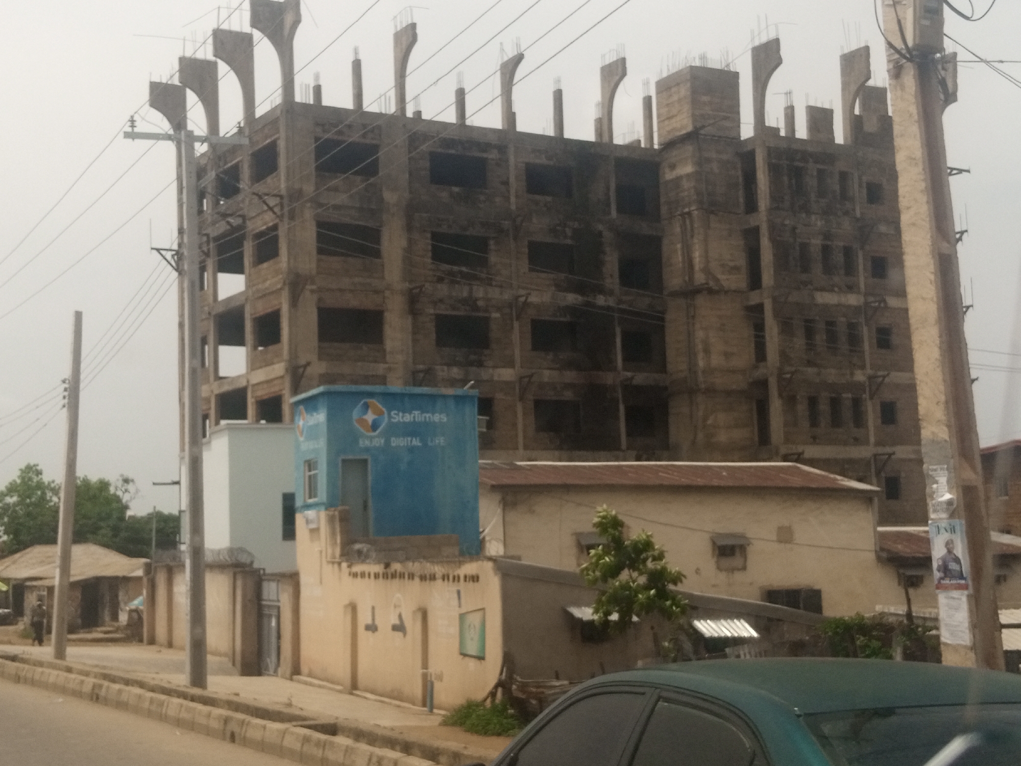 The image depicts an urban scene in Jos, Nigeria, characterized by a multi-story building under construction. The building, made of concrete, has a skeletal appearance with visible rebar on the upper levels. Power lines crisscross the sky, supported by utility poles. In the foreground, a paved road with a concrete curb runs parallel to a sidewalk. Adjacent to the construction site, a smaller, single-story building with a corrugated iron roof is visible. A blue structure, possibly a kiosk or small office, with the "StarTimes" logo and the slogan "ENJOY DIGITAL LIFE," is situated near the construction site. A person, appearing to be a man, is walking on the sidewalk in the distance, near a low-lying building with a thatched roof. The overall atmosphere is overcast, suggesting cloudy weather.