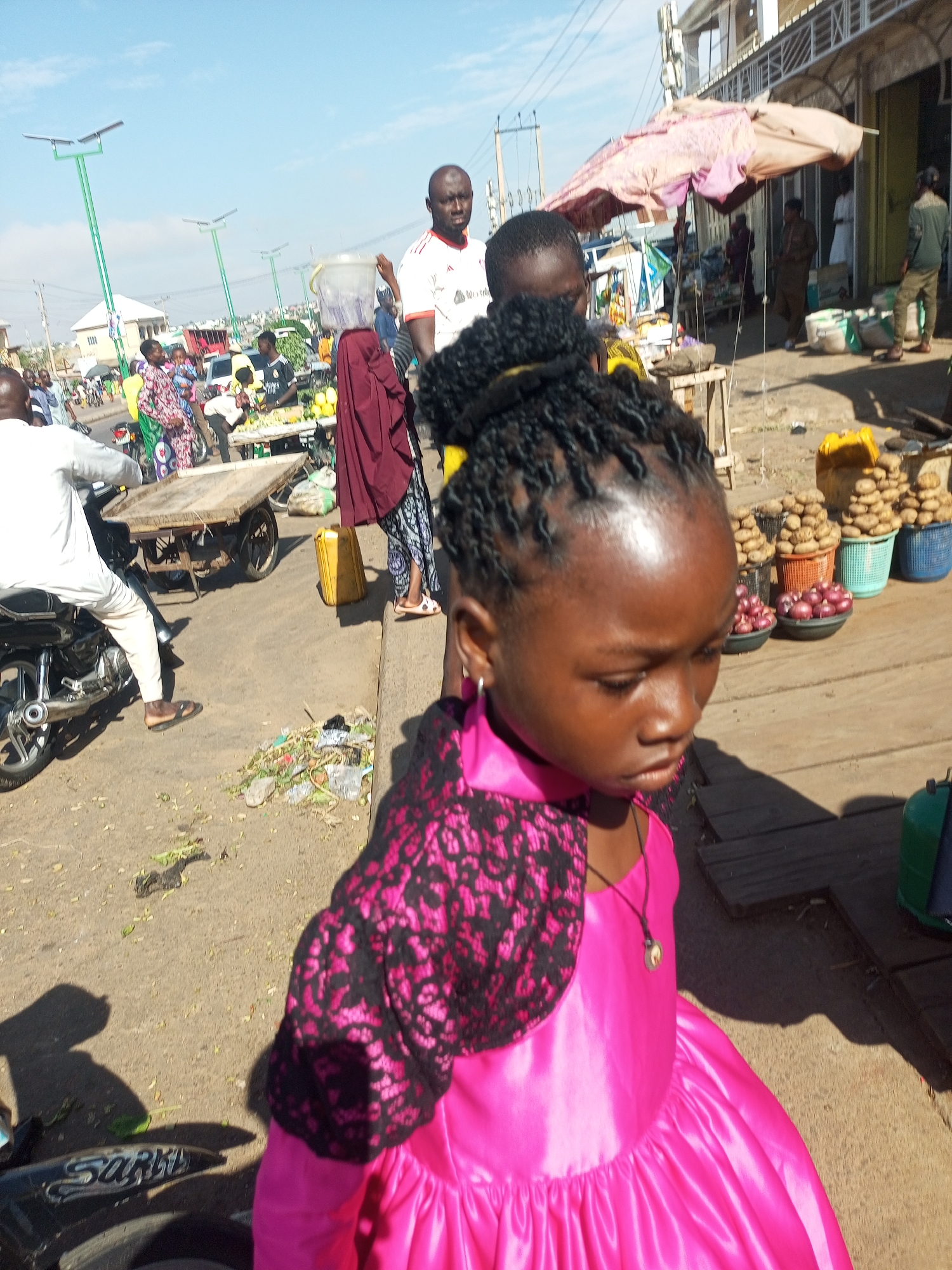 This image depicts a street scene in Tempure, Nigeria, on a bright, sunny day. The foreground is dominated by a young girl wearing a vibrant pink dress with a black lace overlay. Her hair is styled in dark braids. She is looking down and to the right, her expression unreadable. To her left, a motorcycle is partially visible, with the word "Sarki" written on its side. A shadow of a person is cast on the ground to the left of the motorcycle.
The background reveals a bustling market environment. Several people are present, some dressed in traditional attire, others in casual wear. A man in a white shirt with red trim is visible on the left, and another man in a white t-shirt and black shorts stands behind a market stall. Market stalls are laden with produce, including what appear to be potatoes and onions in baskets. A wooden cart is also present. In the distance, solar-powered streetlights are visible, suggesting modern infrastructure within the urban setting. The ground is paved with dirt and some debris is scattered. The overall atmosphere appears lively, typical of a marketplace.