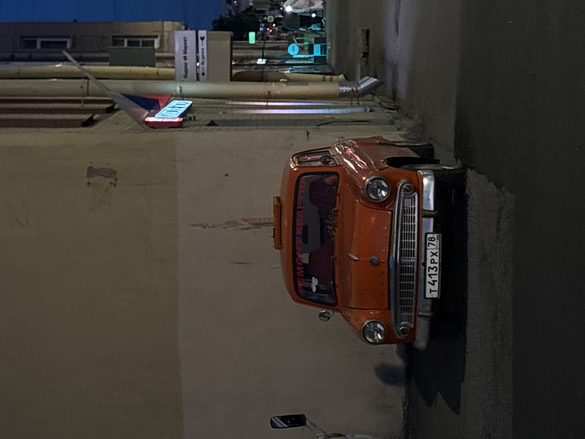 The image depicts an orange vintage Soviet-era car, likely a Moskvich, parked on a street in Saint Petersburg, Russia. The car is the central subject, with its license plate visible: "T 413 PX 78". A pink banner is affixed to the windshield, with Cyrillic text that appears to read "РЕМОНТ КВАРТИР" (Apartment Repair). The car is positioned next to a building with textured beige and light gray walls, showing signs of wear. A metal drainpipe is visible on the building's exterior. To the right of the car, a street scene unfolds, characterized by a sidewalk and road. Several figures are present in the background, appearing as silhouettes due to the low light conditions, suggesting it is nighttime or twilight. A Russian flag is draped from the building. Signs with Cyrillic lettering, including one that reads "Яндекс Маркет" (Yandex Market), are also visible on the building facade. The overall atmosphere is urban and somewhat gritty, with artificial lighting from storefronts and streetlights illuminating the scene. The image captures a moment of urban life in Saint Petersburg, highlighting a piece of automotive history alongside contemporary elements of the city.