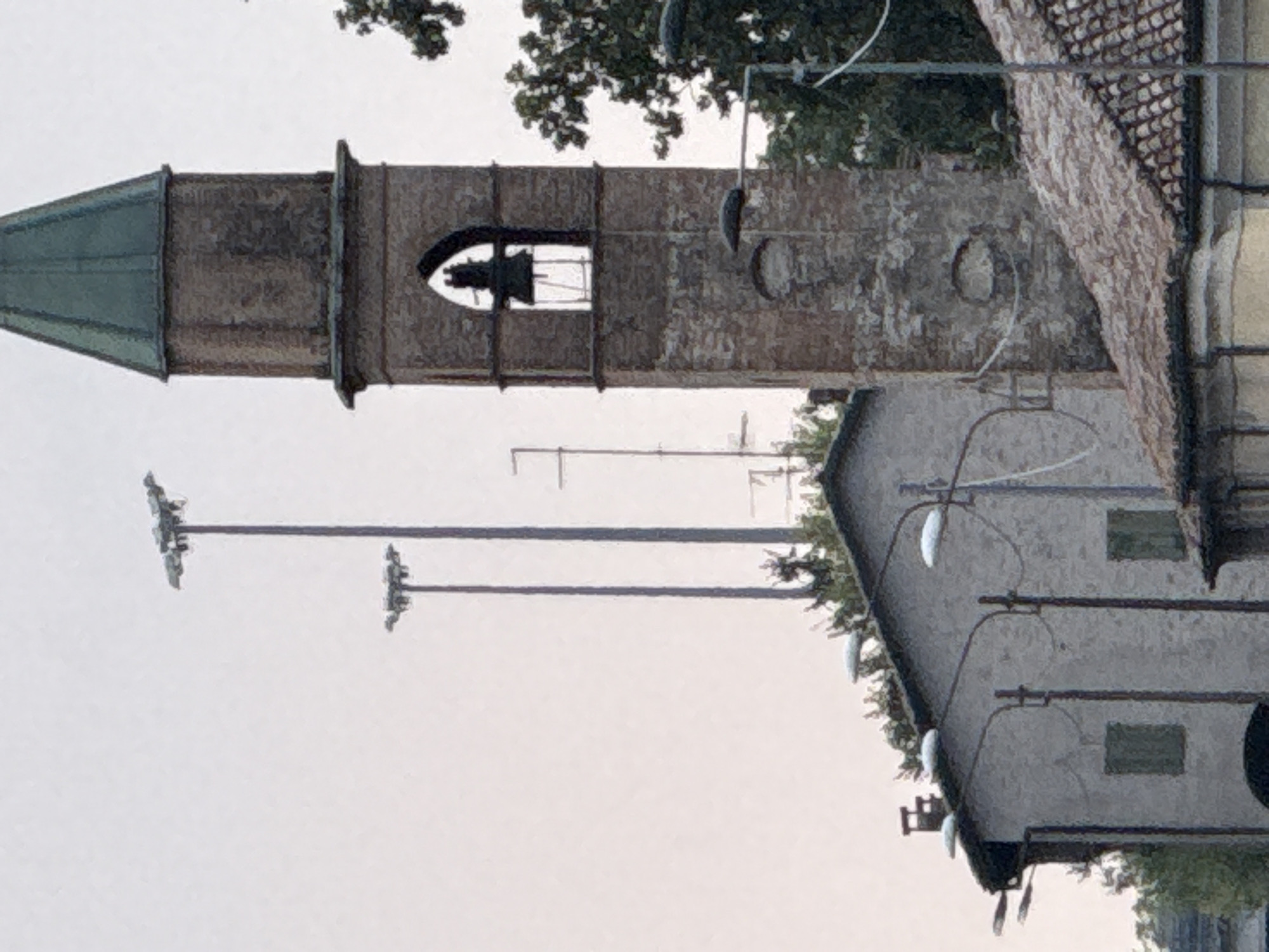 The image depicts a street scene in Fontevivo, Italy, featuring a prominent bell tower with a green, pitched roof. The tower, constructed of aged brick, has an arched opening revealing a bell. To the left of the tower, a light-colored building with a slanted roof and two windows is visible. Multiple street lamps with curved arms are positioned along the street. In the background, two tall utility poles with multiple lights are present. The sky is overcast. The scene appears to be a daytime shot, with no people or animals visible. The overall impression is one of a quiet, possibly historical, urban or semi-urban environment.