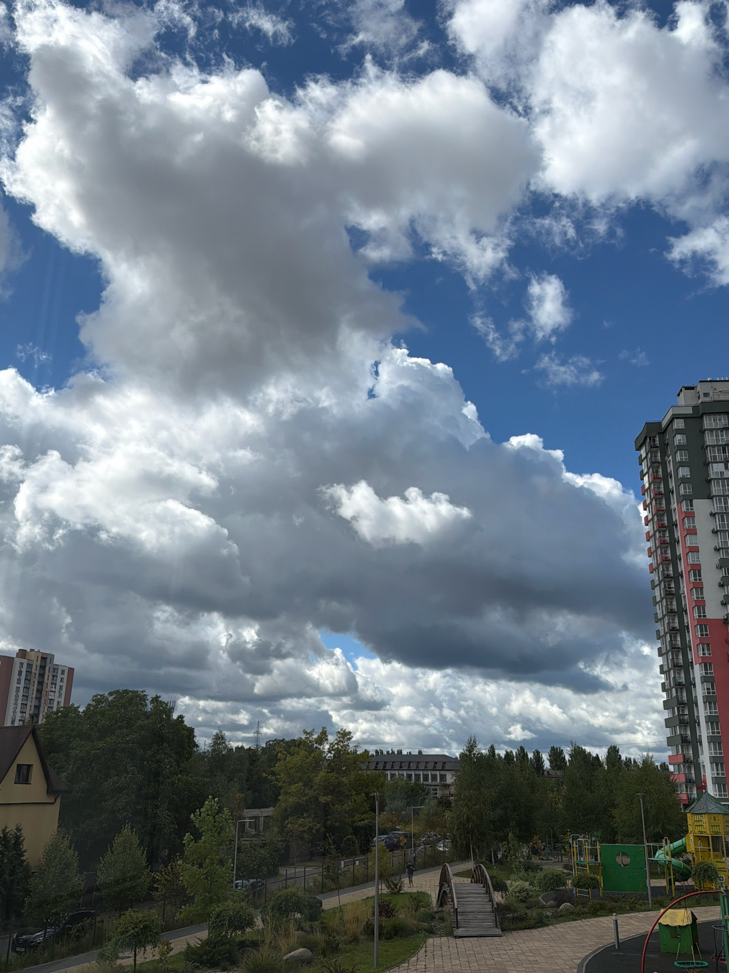 The image captures a cloudy sky over a residential area in Kyiv, Ukraine. In the foreground, a paved walkway leads to a wooden bridge crossing a small waterway, surrounded by greenery and landscaping. To the right, a children's playground with colorful structures is visible. In the middle ground, a dense line of trees separates the foreground from a row of buildings. A tall, modern apartment building with red and green accents stands prominently on the right side of the frame. Another apartment building, lighter in color, is visible in the background on the left. The sky is filled with large, dramatic cumulus clouds, with patches of blue sky peeking through. A person is walking on the path in the lower left quadrant of the image.