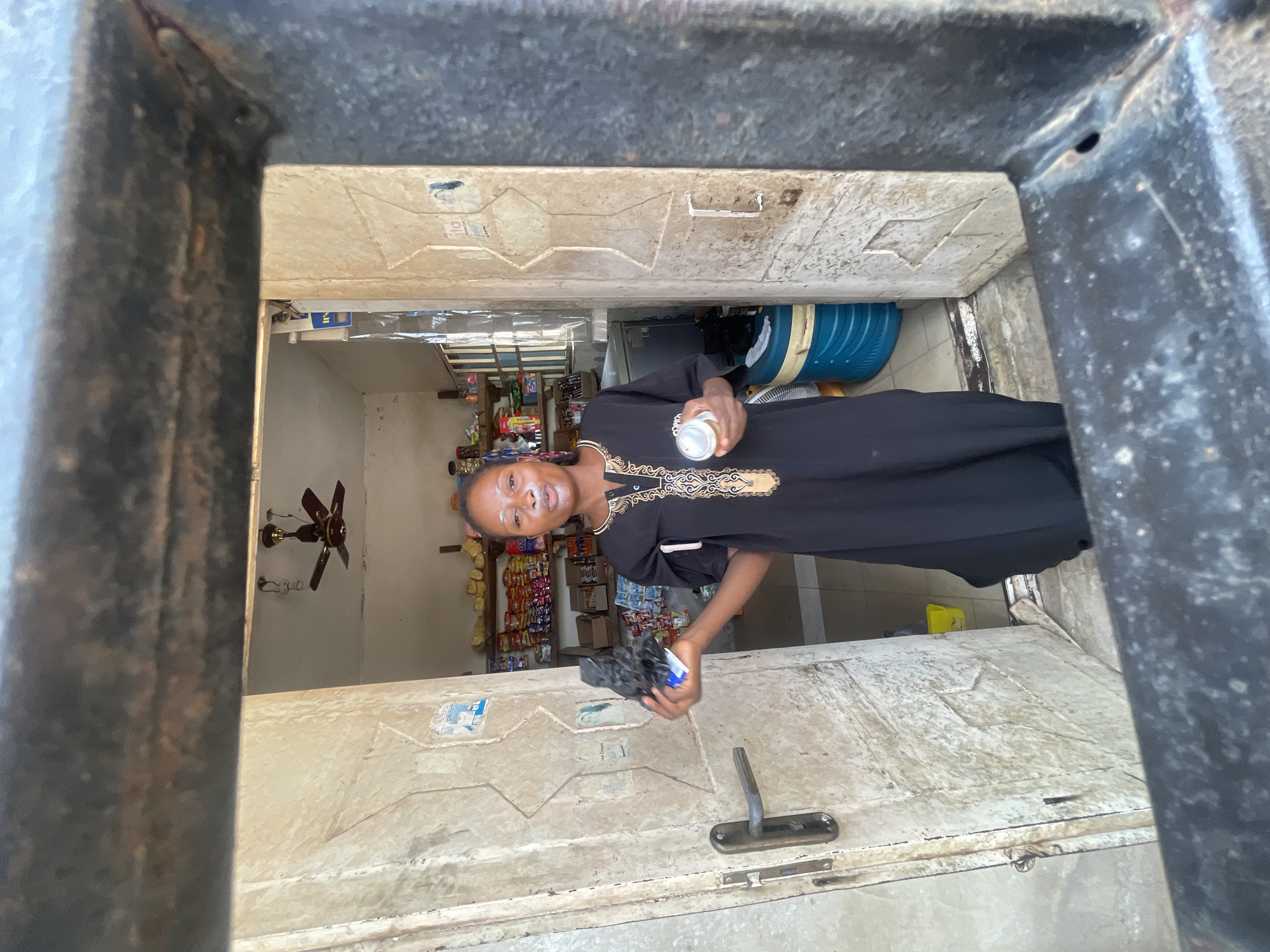 A person, likely a woman, stands framed by a doorway of a store in Lokoja, Nigeria. She is holding a cylindrical container in her right hand and a dark, crumpled object in her left. She wears a black dress with gold-colored embroidery around the neck. The store behind her has shelves stocked with various products, including snack foods, indicating a small convenience or grocery store. A ceiling fan is visible in the upper left portion of the frame. The doorway appears aged and worn. A blue barrel and other items are stacked to the right of the person in the background. The lighting indicates the photo was taken during daylight.