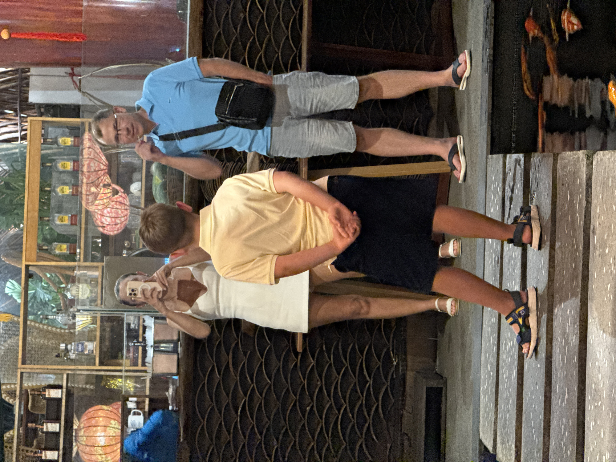 This nighttime image, set in Nha Trang, Vietnam, captures three individuals on what appears to be a walkway or deck alongside a body of water.
In the foreground, a man wearing a light blue polo shirt, grey shorts, glasses, and sandals stands facing slightly left, looking towards the right. A black cross-body bag is slung over his shoulder. Behind him, to his right, another person in a yellow t-shirt and dark shorts has their back to the viewer. Further to the right, a woman in a white dress or top is holding a phone up, seemingly taking a selfie or a picture of something in front of her. Her reflection is visible in the glass surface behind her.
The background reveals an illuminated display cabinet or shop window, showcasing numerous bottles, likely alcohol, arranged on shelves. Decorative elements like a large, reddish-orange spherical lantern and an abstract, intricate light fixture are also visible within the display. The ground beneath the individuals is made of dark planks or stone, reflecting the artificial lights. To the far right, a dark body of water, possibly a canal or river, reflects indistinct lights and shapes. The overall impression is that of an evening outing in a vibrant, commercially active area. The casual attire of shorts and sandals suggests a warm climate. No discernible text is visible in the image.