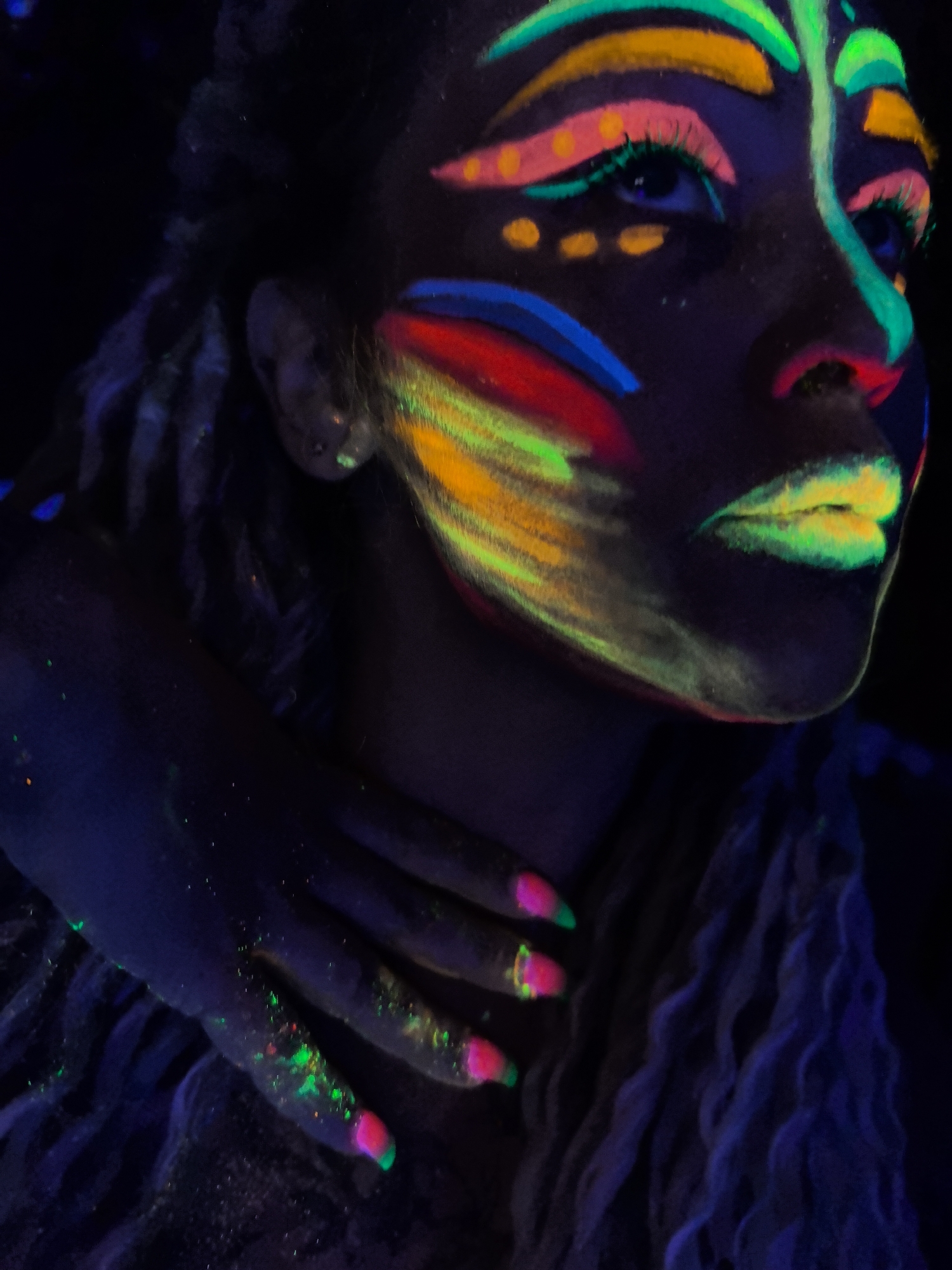 A person is seen in a dark environment with fluorescent paint on their face and body. The paint forms patterns around the eyes, eyebrows, and lips. A hand is placed on the person's neck, with fluorescent paint also on the hand and nails. The person's gaze is directed to the upper right. The person's hair is long and braided. There is no evidence that the image was taken in the city of Noviy, Russia.