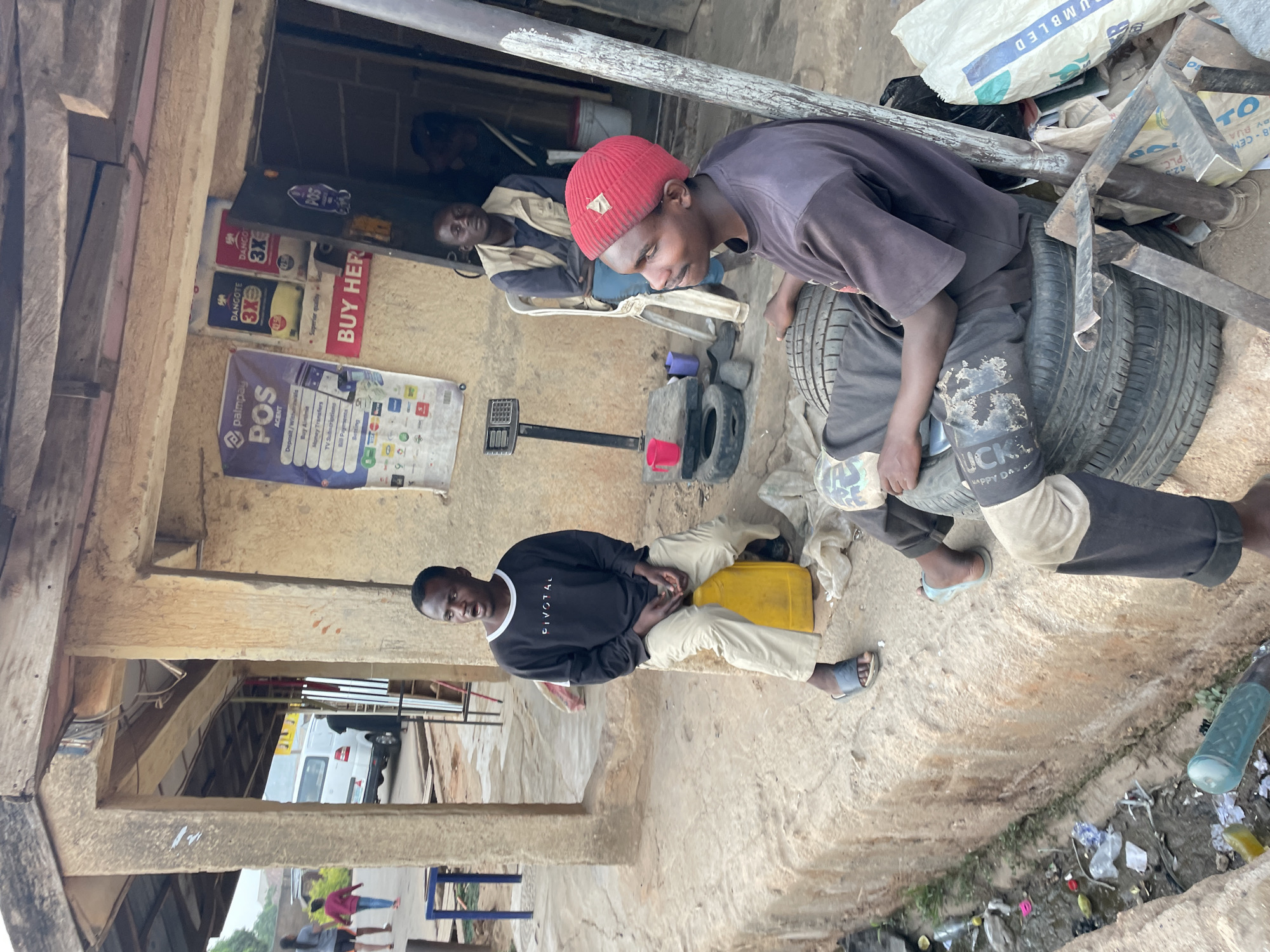 This image depicts an outdoor or semi-sheltered commercial area, likely a repair shop or kiosk, in Jos, Nigeria, during the daytime. The ground is a mix of dirt and concrete, with some litter visible.
Three men are prominent in the foreground and midground. In the immediate foreground, to the right, a man wearing a red beanie and a dark t-shirt is bending over, seemingly inspecting or working with two used car tires stacked on the ground. He holds a small, reflective object in his hand. His trousers are dark, with visible wear or pattern. Next to him are various metal scraps, white sacks (one visible with text), and other debris.
To the left, another man in a black long-sleeve shirt with white trim and light-colored pants sits on the ground, leaning back against a low wall or structure. A yellow jerrycan is placed beside him. Further into the background, inside what appears to be a small, open-fronted structure, a third man in a light and dark jacket is seated on a white plastic chair, looking towards the foreground.
Signs are affixed to the structure's walls: a blue and white "palmpay POS AGENT" banner with services like "Deposit / Withdrawals," "Buy Airtime," "Money Transfers," "TV Subscriptions," and "Bill Payments." Another sign, red and white, reads "BUY HER." Below that, a blue and white "DANGOTE 3X" sign, possibly for cement, is visible, with "Superis" and "quality" partially visible.
The overall scene suggests a bustling, functional outdoor workspace. Visible text includes: "PIVOTAL," "palmpay POS AGENT," "Deposit/Withdrawals," "Buy Airtime," "Money Transfers," "TV Subscriptions," "Bill Payments," "DANGOTE 3X," "Superis," "BUY HER," "HAPPY DA," "UCKL," "UMBLED," "42.5 PLC.," "BY BUA," and "TO."