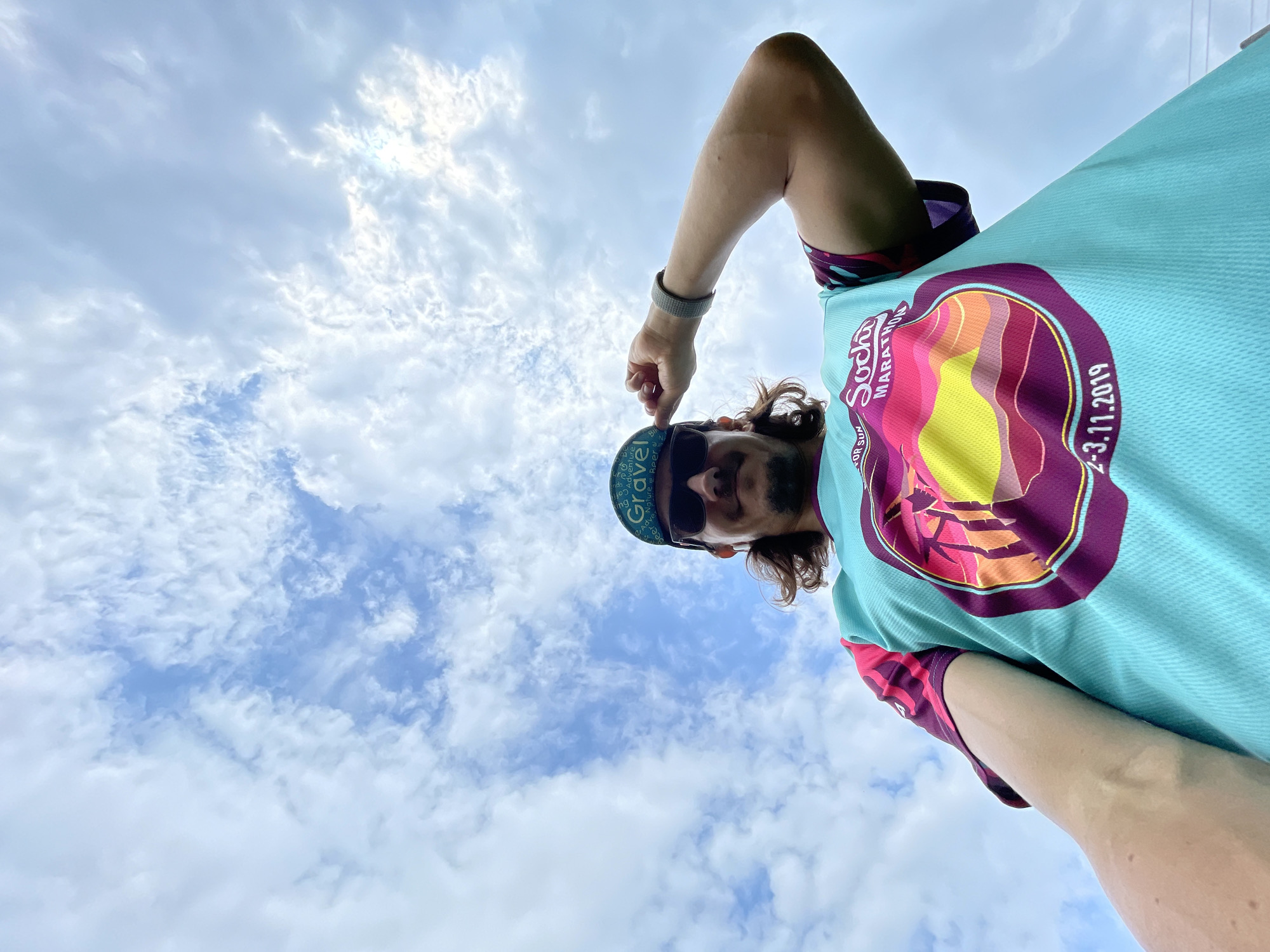 A low-angle, wide shot captures a person from the chest up, posing under a cloudy sky. The person, wearing sunglasses and a teal athletic shirt with a colorful circular graphic on it, is holding the brim of a green baseball cap. The graphic on the shirt is a sunset with palm trees and text that reads "Sochi Marathon" and "2-3.11.2019." The person has long, dark hair and a mustache. The sky is filled with fluffy white clouds against a bright blue background. The person is outdoors, with the camera pointed upward towards the sky.