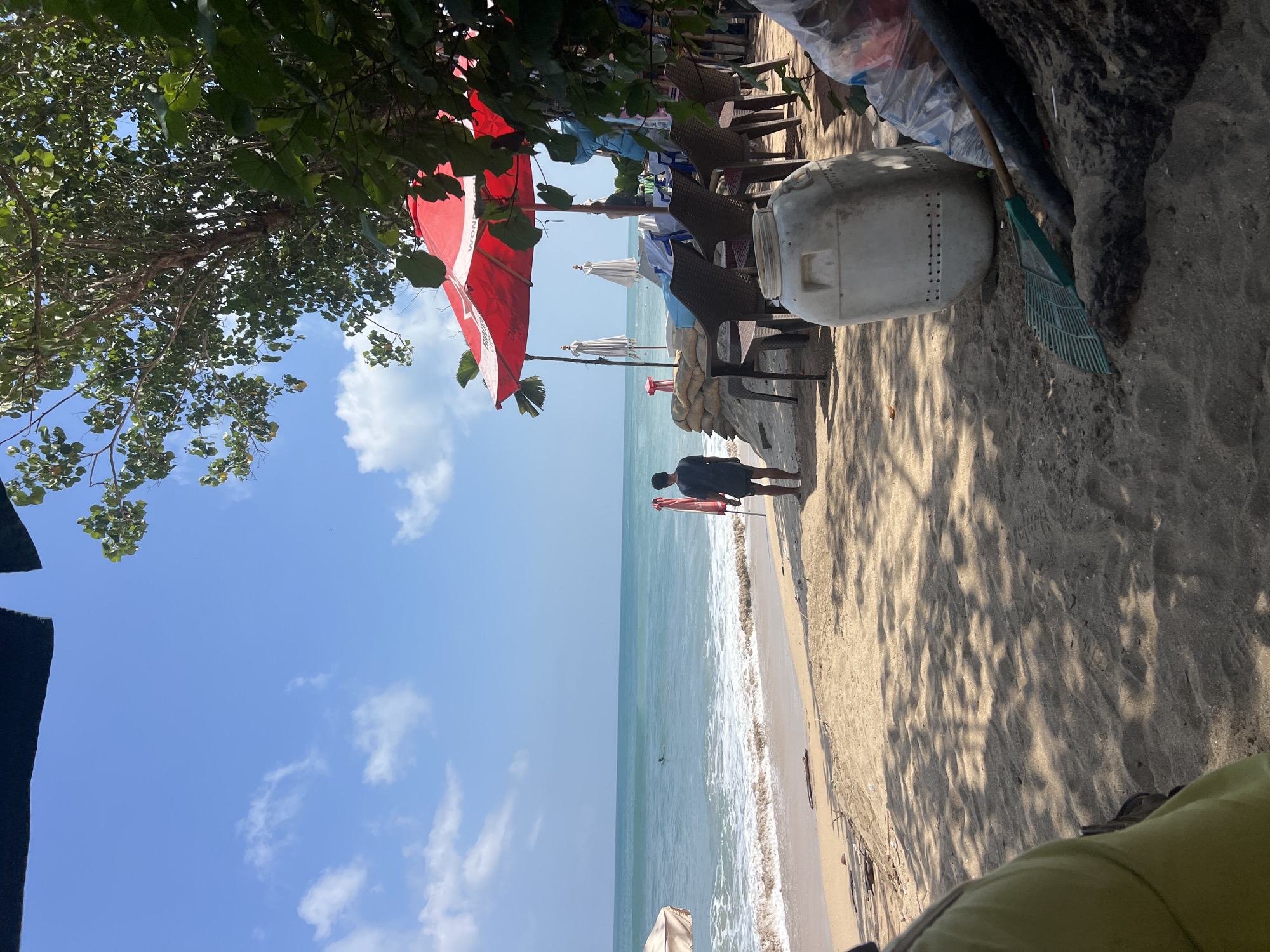 A wide shot captures a sandy beach scene in Kuta, Indonesia, under a bright blue sky with scattered white clouds. In the foreground, the sandy beach is dotted with shadows cast by unseen trees. A large, light-colored plastic container sits on the sand, alongside a green rake and a black pipe leaning against a mound of dark material, possibly dirt or debris. A person wearing a green garment is partially visible in the lower left corner, facing away from the camera. Further back, a man in a dark t-shirt and shorts stands on the sand, facing the ocean.
The middle ground features the shoreline where gentle waves are breaking onto the sand. In the distance, the turquoise ocean stretches to the horizon. Two red and white beach umbrellas are prominent features, one fully open and another partially folded. Behind them, several plastic chairs are arranged around tables, suggesting a seating area for patrons. Sandbags are stacked near the umbrellas, possibly as a barrier or support. A person is visible in the ocean, swimming or wading. The scene depicts a relaxed, beachside atmosphere, likely a tourist area in Kuta.