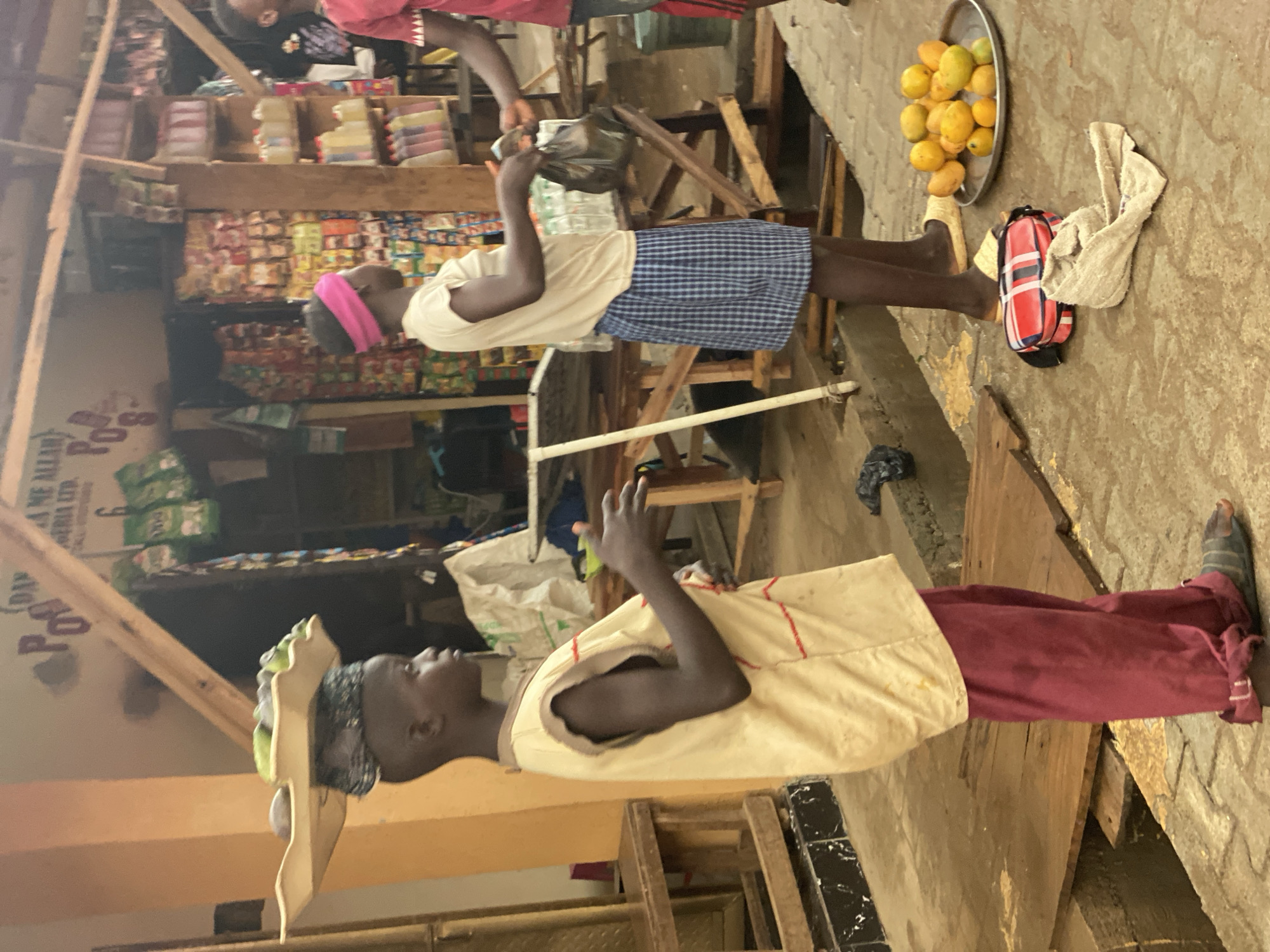 The image depicts a scene at a market in Kafanchan B, Nigeria. There are at least two individuals present, presumably customers or vendors. One person is wearing a white shirt and blue checkered shorts, standing in a partially obstructed area. Another person is wearing a tan shirt with red pants. A metal tray containing mangoes rests on the ground next to a red and white striped bag. The setting appears to be a market, with shelves stacked with goods, visible in the background. The overall environment is bustling and informal.