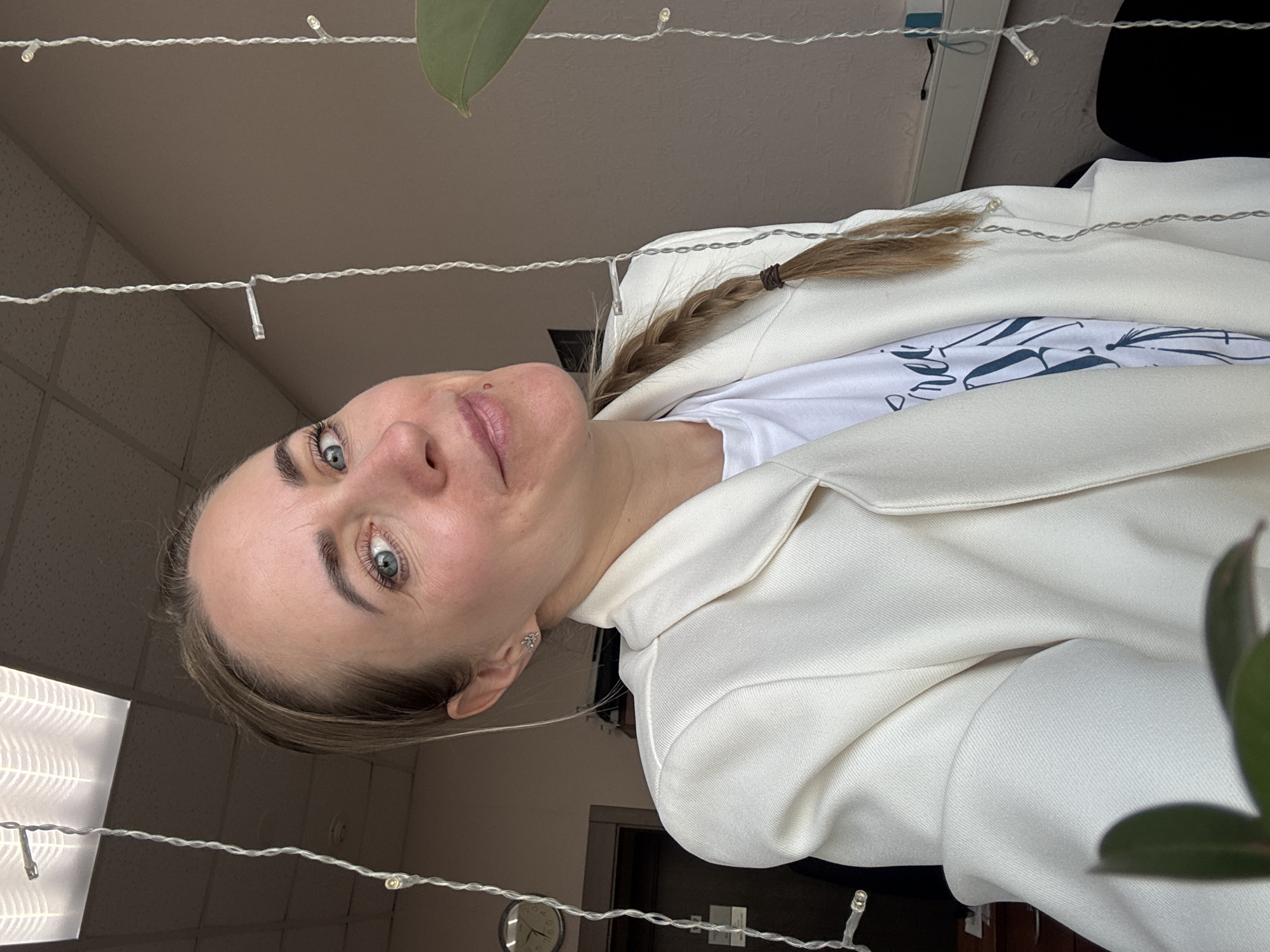 A woman with fair skin and blue eyes, wearing a white blazer over a white t-shirt, looks directly at the camera. She has a single braid pulled to the side. The scene is indoors. Background details include string lights, a leaf, a clock, and what appears to be an office setting. There is no specific, verifiable information to confirm it is located in Chaikovsky, Russia.
