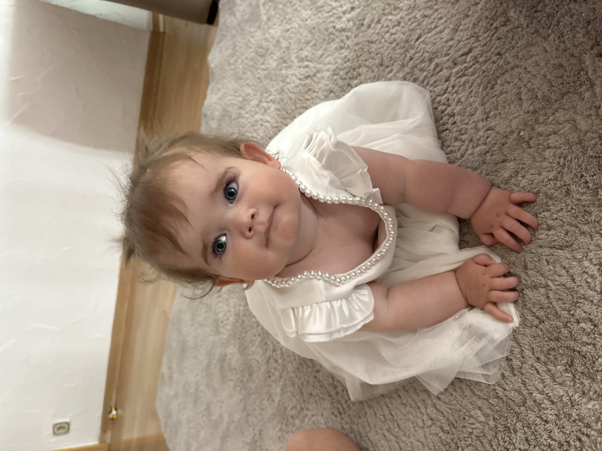 A baby wearing a white dress with pearl trim is lying on a beige shag rug. The baby has blue eyes and light brown hair. The background includes a white wall and a light brown wooden element. The location cannot be determined from this image.