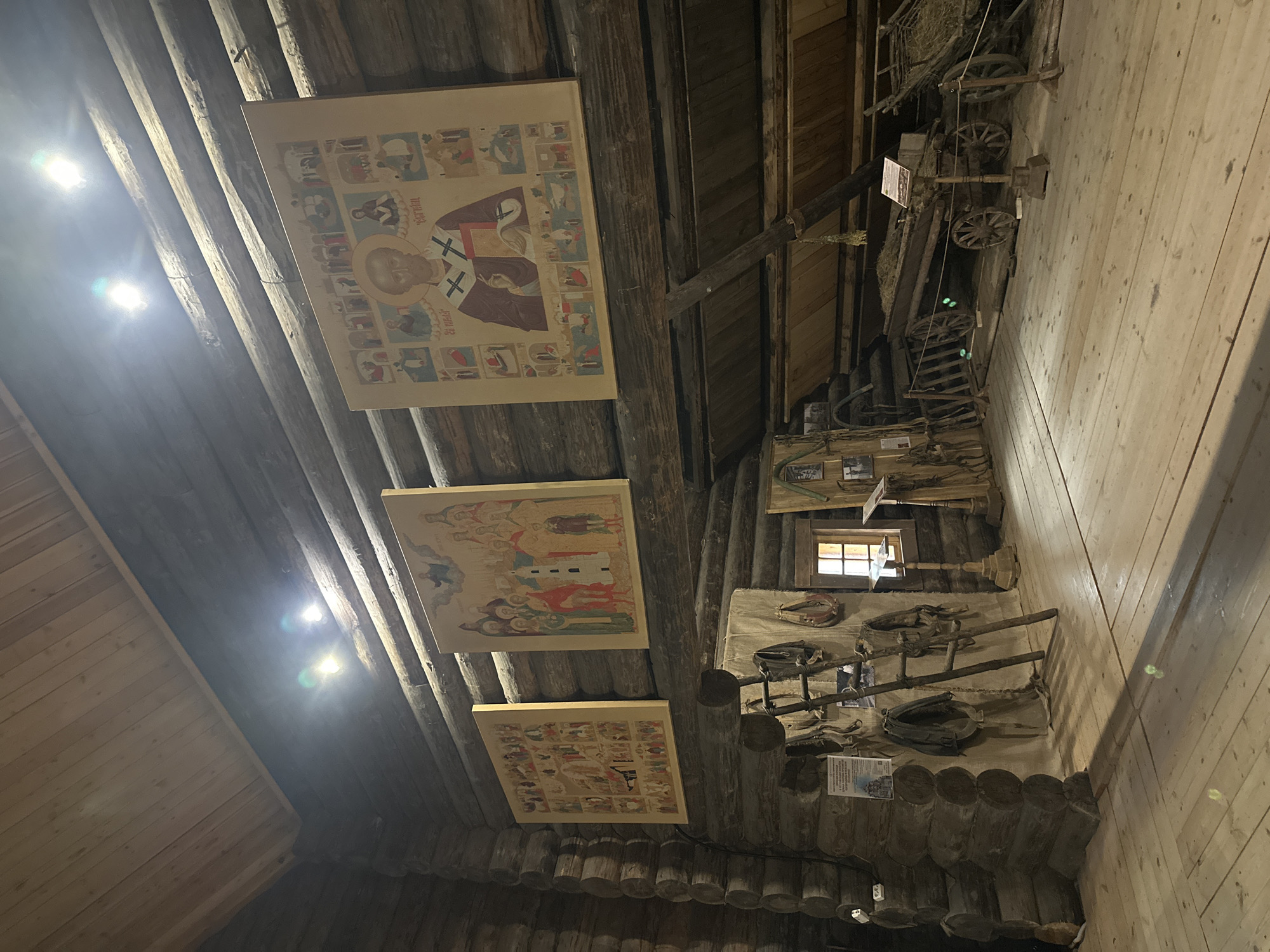The image depicts the interior of a rustic wooden structure, likely a museum exhibit, located in Nevskii Parkleskhoz, Russia. The walls and ceiling are constructed from rough-hewn logs. Three large, framed religious icons hang on the wall. The icon on the far right depicts Saint Nicholas surrounded by smaller scenes. The middle icon shows a gathering of figures, and the leftmost icon is also a religious scene with multiple figures.
In the foreground, various agricultural tools and equipment are displayed. A wooden ladder leans against a wall, and harnesses and other leather goods are hung on the wall and displayed on a canvas. A small window with wooden shutters is visible. Further into the space, two horse-drawn carts are parked, one filled with hay. The floor is made of wooden planks. The lighting appears to be artificial, with several bright spotlights mounted on the ceiling, casting distinct beams and reflections on the wooden surfaces. No people are visible in the image.