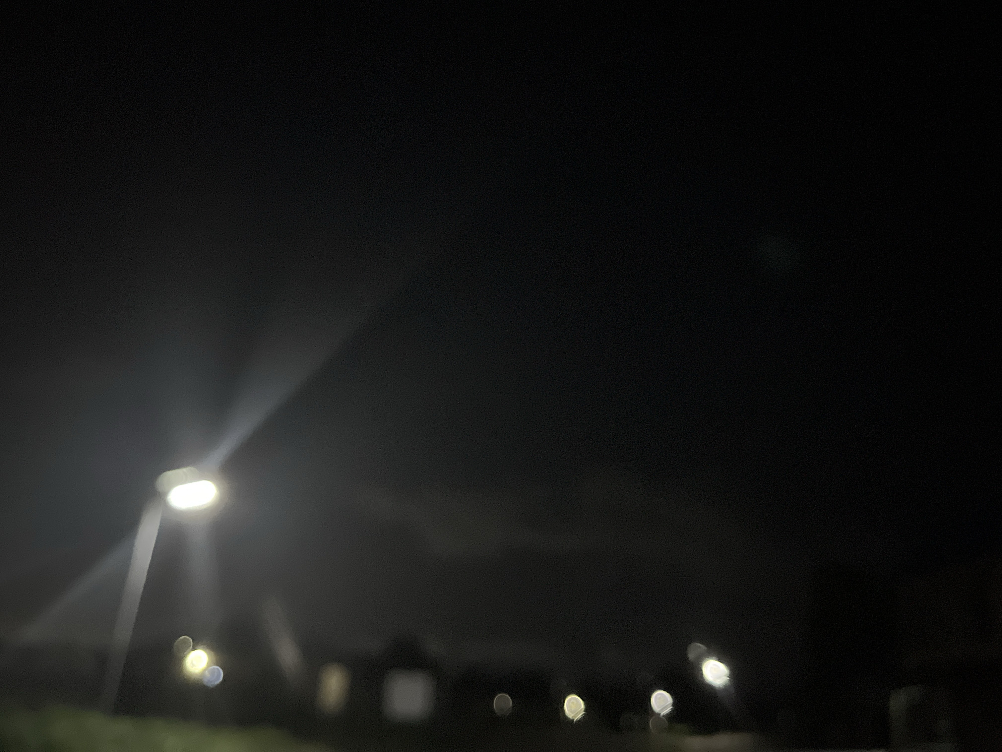 The image depicts a nighttime scene in an unknown city. There are multiple streetlights illuminating the scene, some of which are out of focus. In the background, there are dark shapes suggesting buildings or trees. The environment appears to be urban or suburban, with the focus predominantly on the artificial lighting sources. No people are visible.