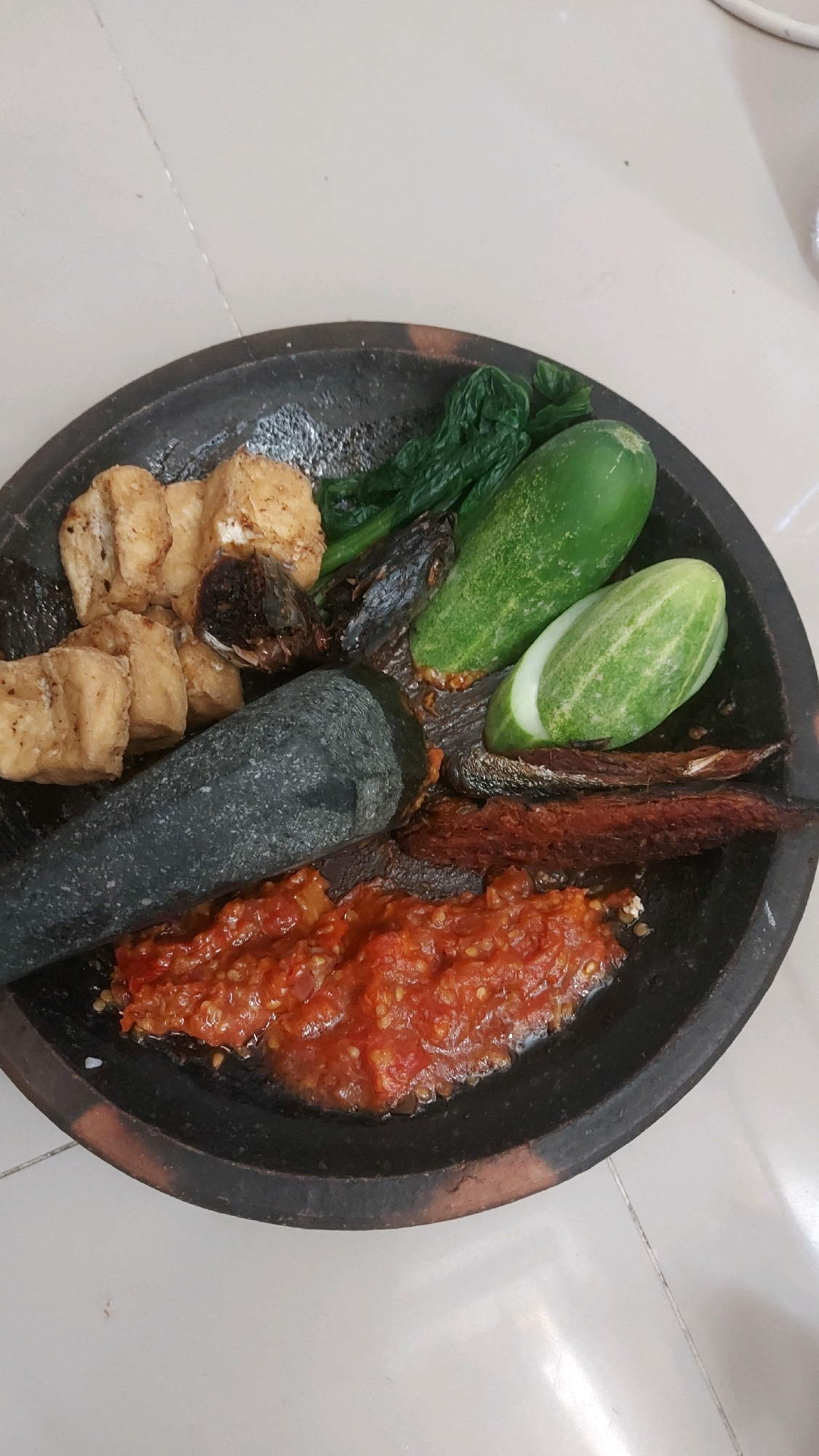 A mortar and pestle are placed on a dark, circular stone plate. The plate contains a variety of food items. In the foreground, there is a red chili paste. To the left of the chili paste, a dark gray, stone pestle rests horizontally. To the left of the pestle are several pieces of fried tofu. Behind the tofu and pestle are some dried fish and a bunch of cooked green leafy vegetables. To the right of the vegetables are two halves of a cucumber, one whole and one sliced. The entire plate is positioned on a light-colored tiled floor. The setting is a home kitchen in Sedati Gede, Indonesia.