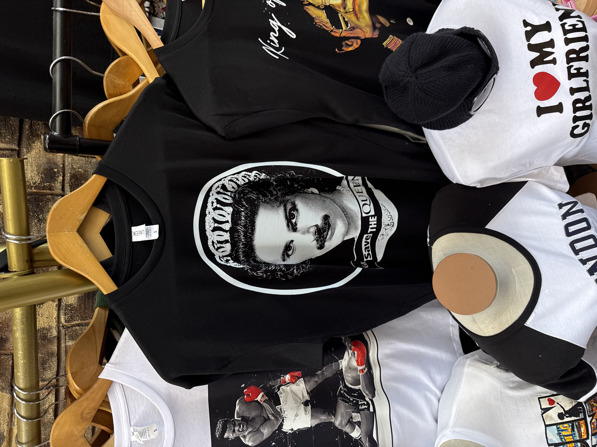 A pile of T-shirts is hung on wooden hangers on a metal rack. The shirts are of different designs: one has a black and white image of Freddie Mercury with "Save The Queen" text; another has a black and white image of boxers, and another reads "I <3 MY GIRLFRIEND". A black beanie and a display head are placed among the shirts. The location is a city of Unknown, Unknown.