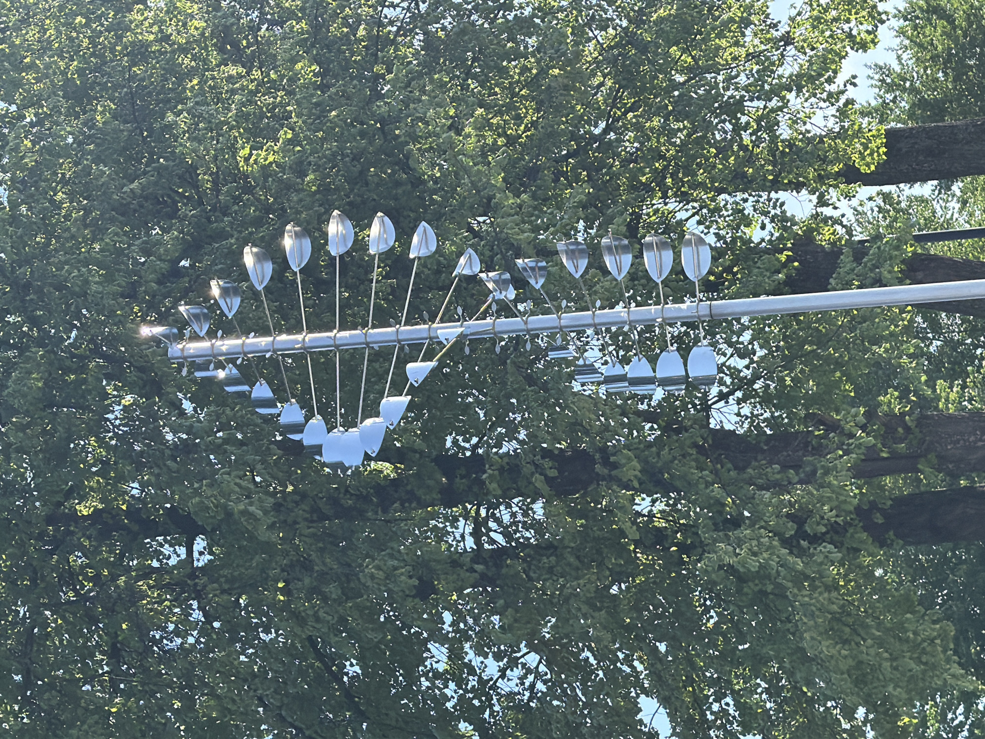 The image shows a horizontal anemometer with a silver metal frame and numerous silver metal cups rotating in the wind. The anemometer is mounted on a pole against a background of green trees and blue sky. The location is not identifiable as Votkinsk, Russia.