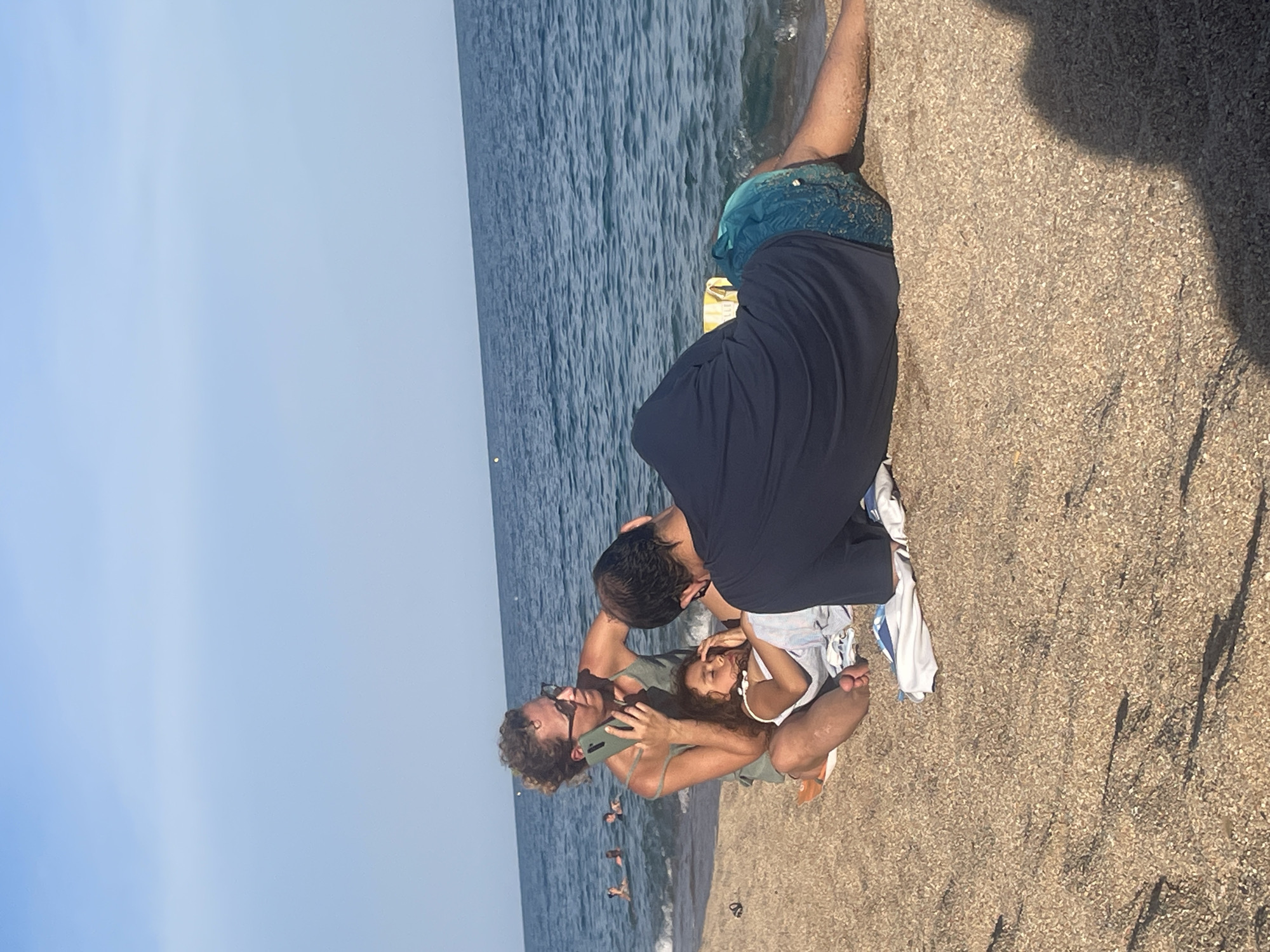 In Canet De Mar, Spain, on a sandy beach, a woman with curly hair and sunglasses is seated on the sand, holding a smartphone to her ear and talking. A young girl, lying on her side with her eyes closed and appearing to be asleep or resting, is positioned on the woman's lap. A man, wearing blue patterned swim trunks and a dark blue t-shirt or rash guard, is lying on his stomach next to them, facing away from the camera. The sea is visible in the background with gentle waves and a clear blue sky overhead. Some other people are in the water further offshore. The sand is a light brown color with small pebbles and some shadows cast across it.