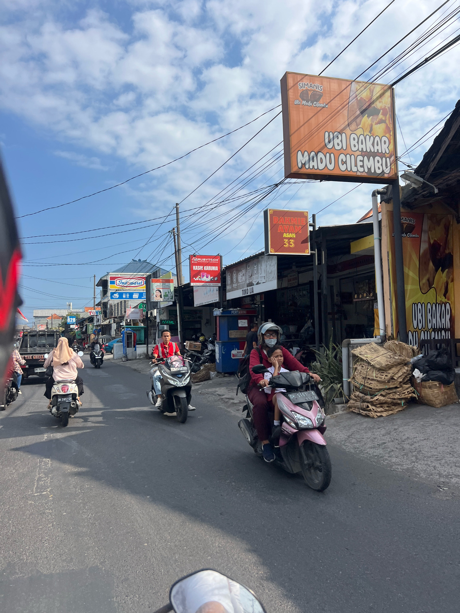 In Denpasar, Indonesia, a street scene unfolds under a partially cloudy sky. The foreground is dominated by a gray asphalt road, with the lower right corner showing a white rearview mirror, indicating the perspective of a vehicle. Several motorcycles navigate the road. On the left, a woman in a pink hijab rides a white scooter, with a person behind her. Further ahead, a man in a red shirt is on a gray scooter, accompanied by a child in the front. The child, wearing a pink shirt and dark pants, looks to the right. The background features various establishments, including a sign for "Indomaret," a convenience store, and other businesses. Prominent signage includes an orange banner advertising "UBI BAKAR MADU CILEMBU" (Grilled Honey Cilembu Sweet Potato) and another red sign for "BAKMIE AYAM ASAK 33" (Chicken Noodles Cooked 33). Stacks of woven mats and black trash bags are visible on the right side of the street, beside one of the shops. The scene captures a typical urban environment with bustling traffic and commercial activity.