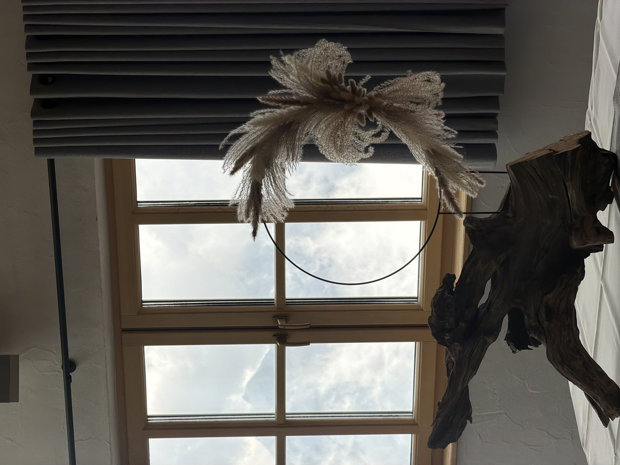 The image depicts an indoor scene with a window providing a view of a cloudy sky and a distant mountain. In the foreground, a piece of driftwood rests on a white tablecloth. To the right of the driftwood, a decorative arrangement of dried pampas grass is suspended by a thin black hoop, positioned in front of a dark grey curtain. The window has wooden frames and appears to be a casement window with two handles. The wall on the left has a textured finish. The scene appears to be set within a dwelling or commercial space in Garmisch-Partenkirchen, Germany, given the architectural style and natural backdrop visible through the window. There are no people or animals present.