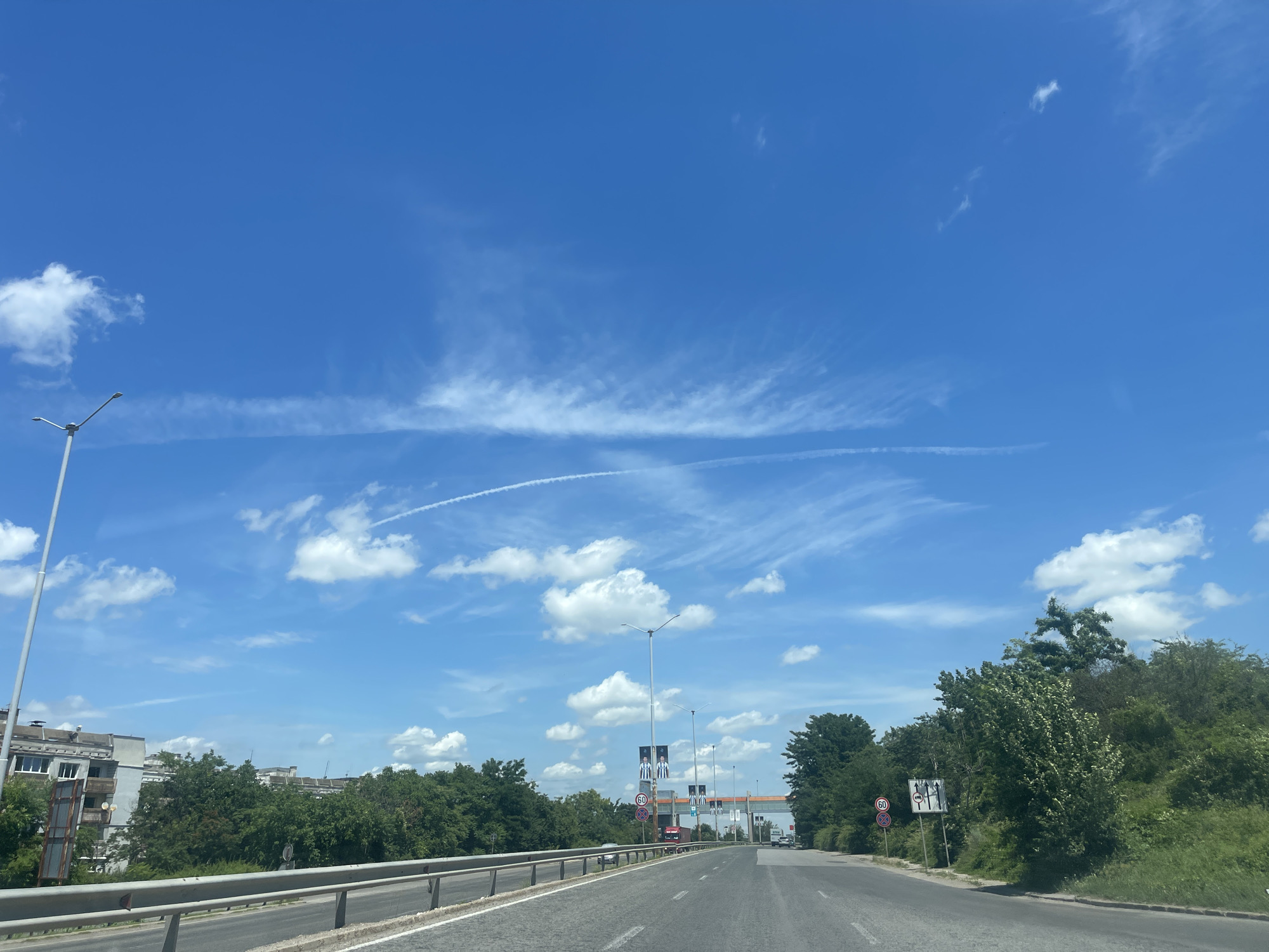 The image depicts a clear blue sky with scattered white clouds and contrails from an airplane. The scene appears to be on a highway or a major road within or on the outskirts of Ruse, Bulgaria. On the left side of the road, there are several multi-story apartment buildings. Trees and dense greenery line the right side of the road and cover a hillside. There are streetlights and several road signs visible, including speed limit signs and directional signs. There are no people or vehicles clearly visible in the immediate foreground, but some vehicles can be seen further down the road in the distance. The overall impression is a bright, sunny day in a semi-urban or suburban environment.